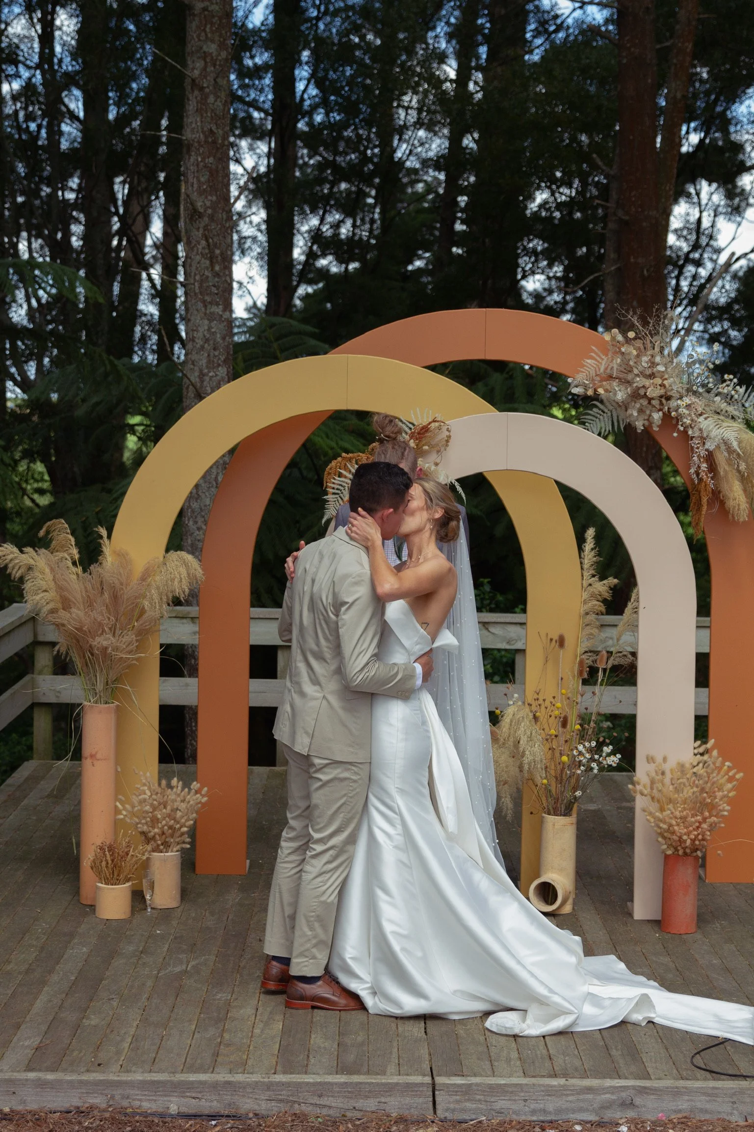 A couple in wedding attire sharing a kiss at an outdoor wedding ceremony with a modern, colorful arch and dried floral decorations.  Wedding Photography Waikato New Zealand. Wedding Photography Hamilton New Zealand. Northland Wedding Photography