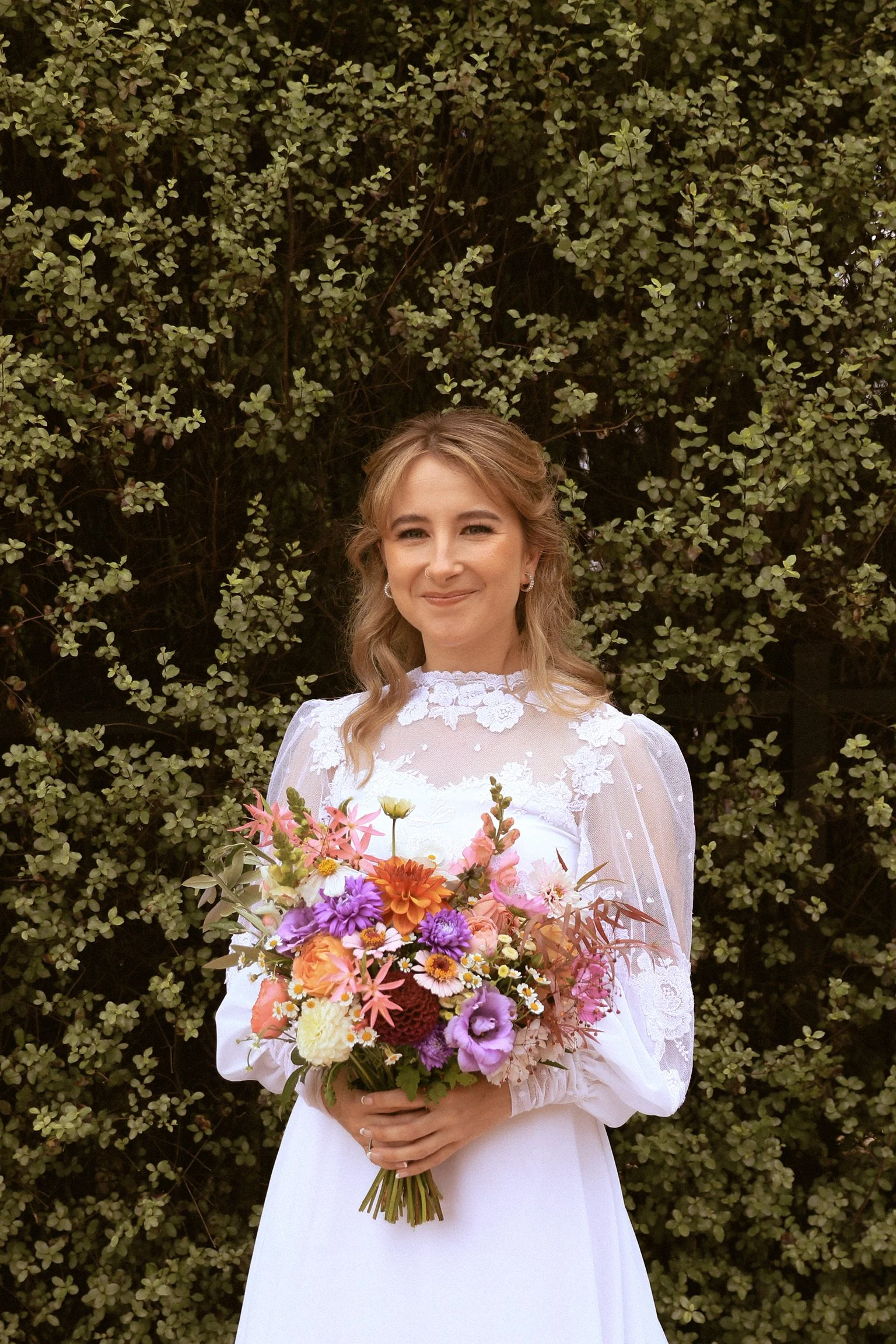 A woman in a white dress holding a colorful bouquet of flowers, standing in front of a leafy green background. Wedding Photography Waikato New Zealand. Wedding Photography Hamilton New Zealand