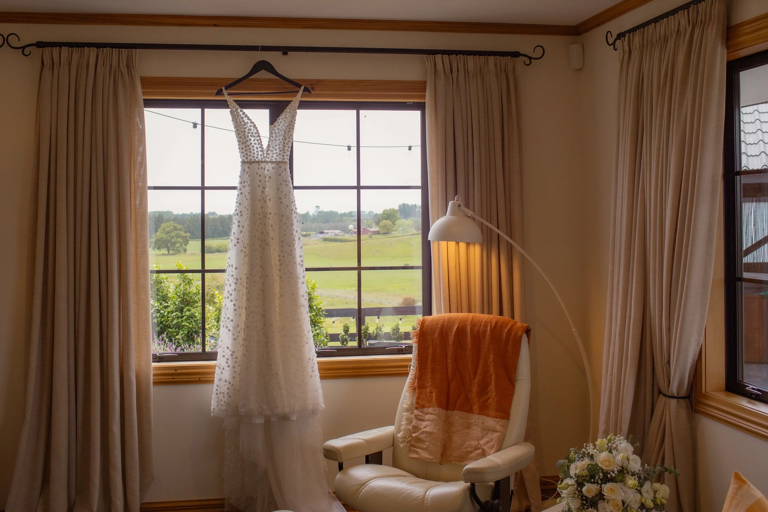 A white wedding dress hanging on a black hanger in front of a window with an outdoor view. The room has beige curtains, a white chair with an orange towel draped over it, a floor lamp, and a bouquet of white roses on a table.