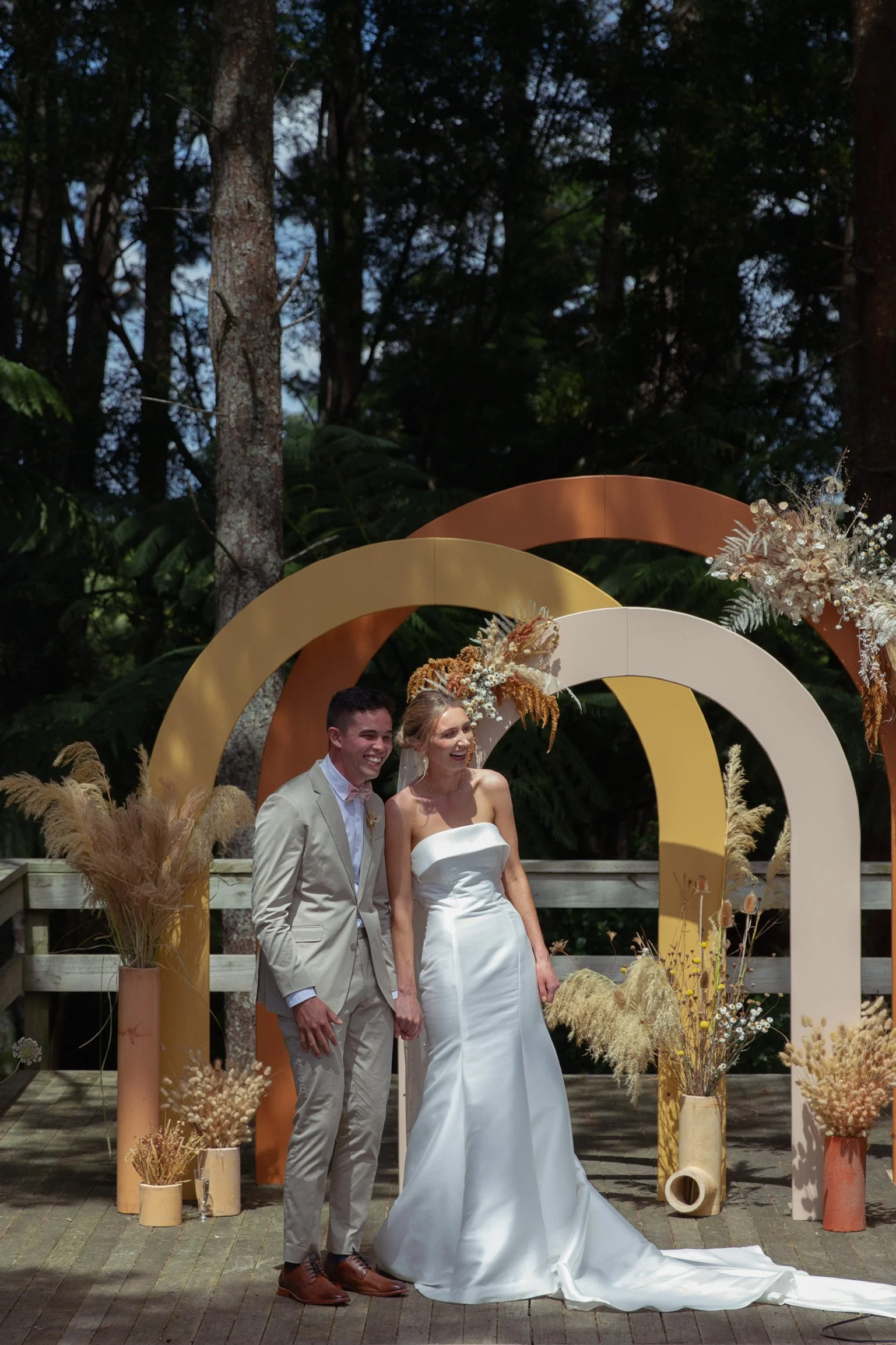 A newlywed couple, standing together outdoors on a wedding day, smiling. The bride is wearing a strapless white wedding dress, and the groom is in a beige suit. Behind them is a decorative arch decorated with dried floral arrangements and surrounded 