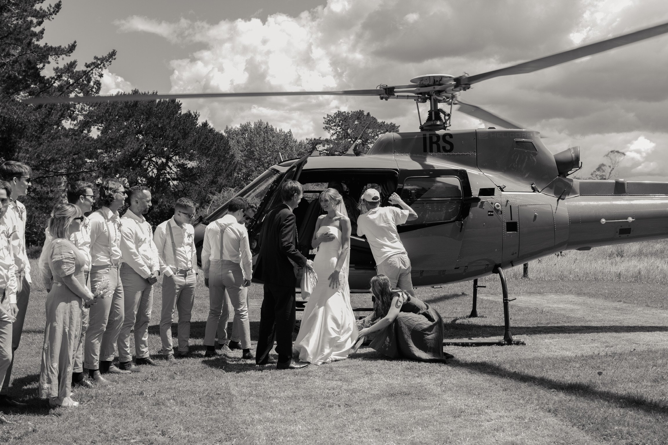 A bride and groom in wedding attire standing beside a helicopter on a grassy field, surrounded by wedding guests, with a woman adjusting the bride's dress.  Wedding Photography Waikato New Zealand. Wedding Photography Hamilton New Zealand. 