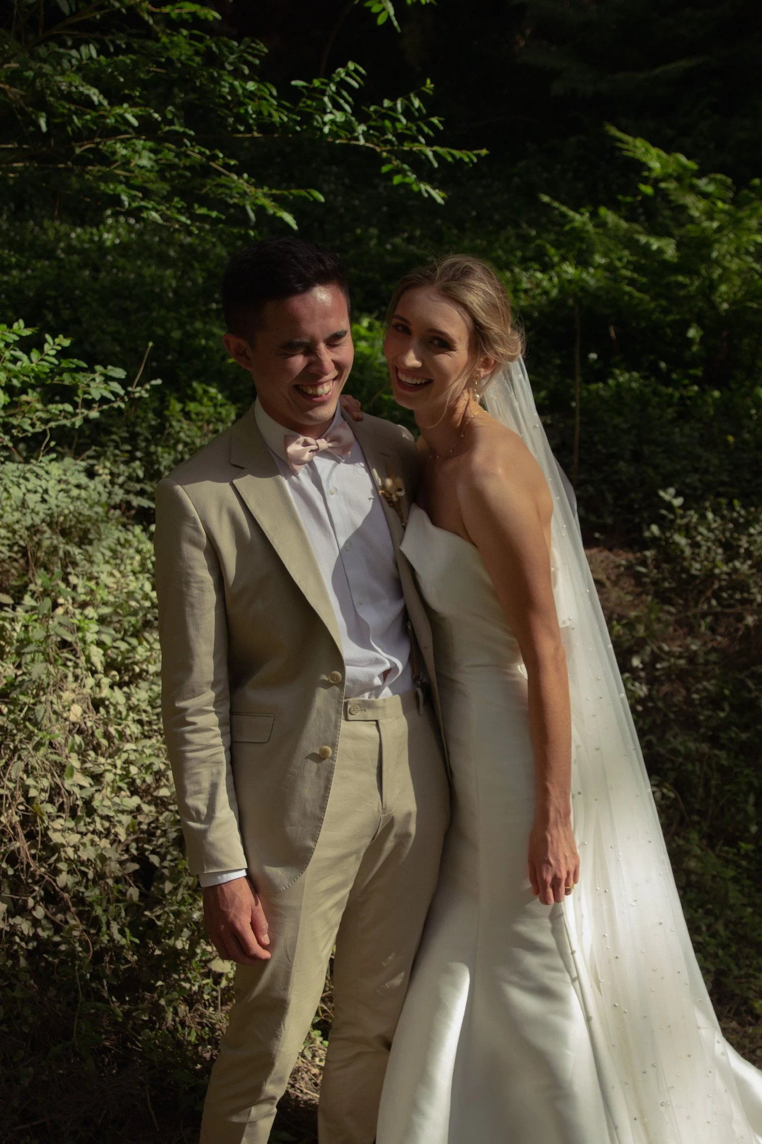 A bride in a white wedding gown and a groom in a beige suit with a bow tie standing outdoors in a wooded area, smiling and laughing.  Wedding Photography Waikato New Zealand. Wedding Photography Hamilton New Zealand. Northland Wedding Photography