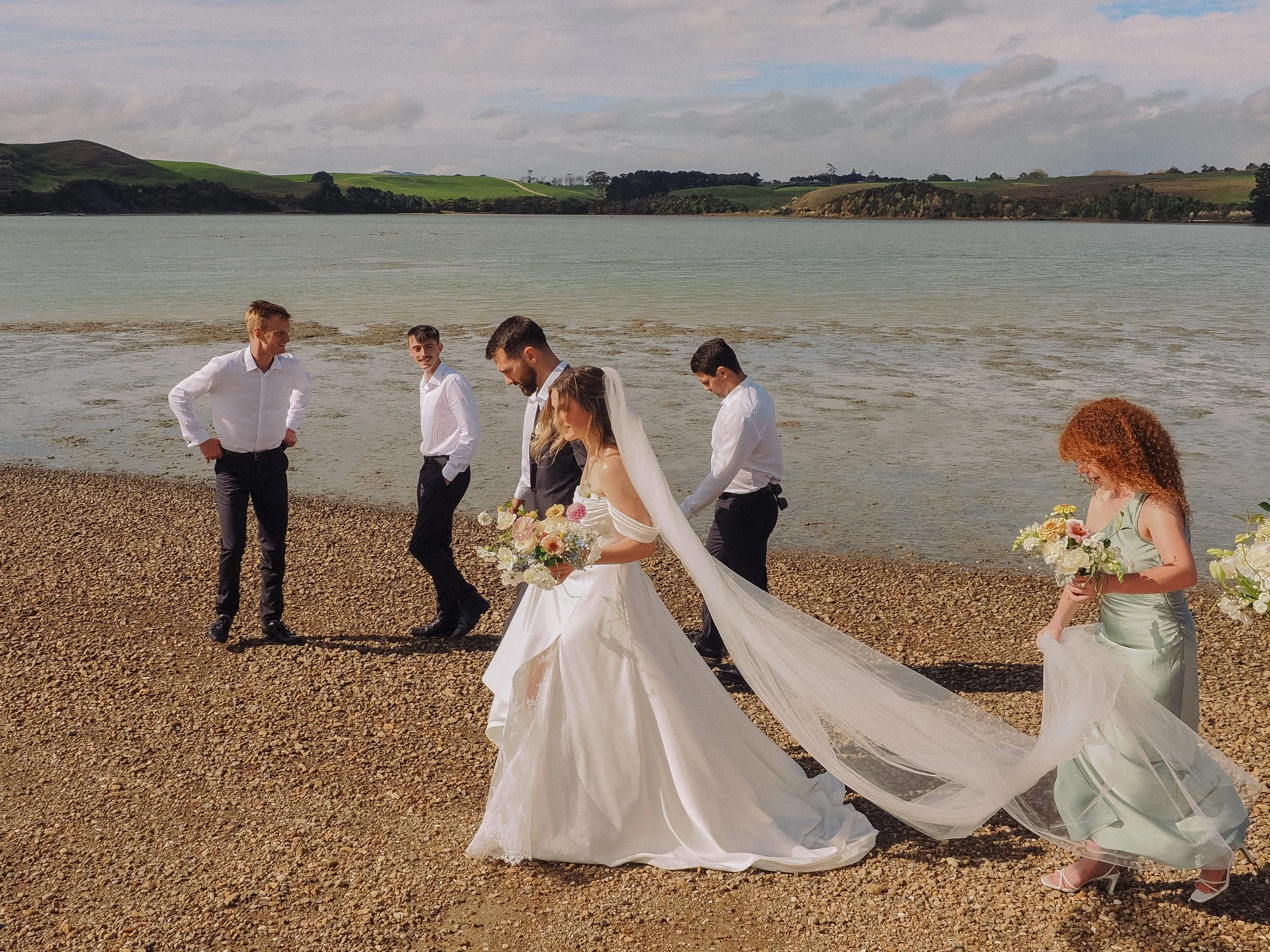 A wedding party with a bride in a wedding dress holding a bouquet of flowers, a groom in a suit, and four attendants by a lakeshore with hills and cloudy sky in the background.  Wedding Photography Waikato New Zealand. Wedding Photography Hamilton 