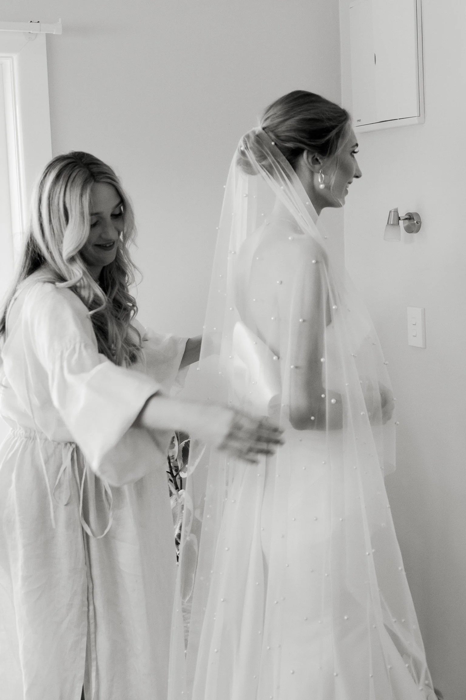 A bride in a wedding dress with a veil, smiling and standing in a room while another woman helps her prepare.  Wedding Photography Waikato New Zealand. Wedding Photography Hamilton New Zealand. Northland Wedding Photography