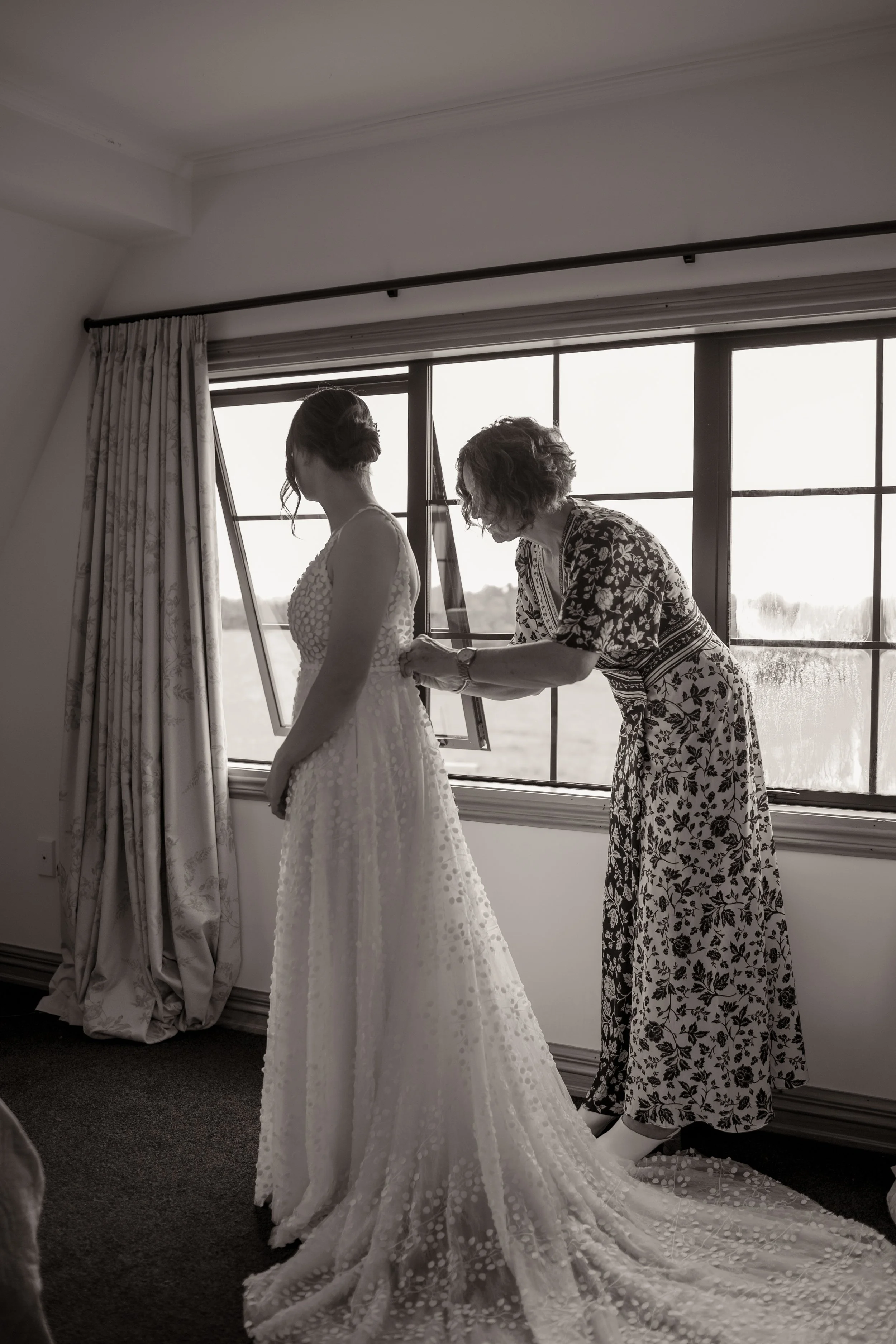 A woman helping a bride adjust her wedding dress inside a room with large windows and curtains.  Wedding Photography Waikato New Zealand. Wedding Photography Hamilton New Zealand. Northland Wedding Photography