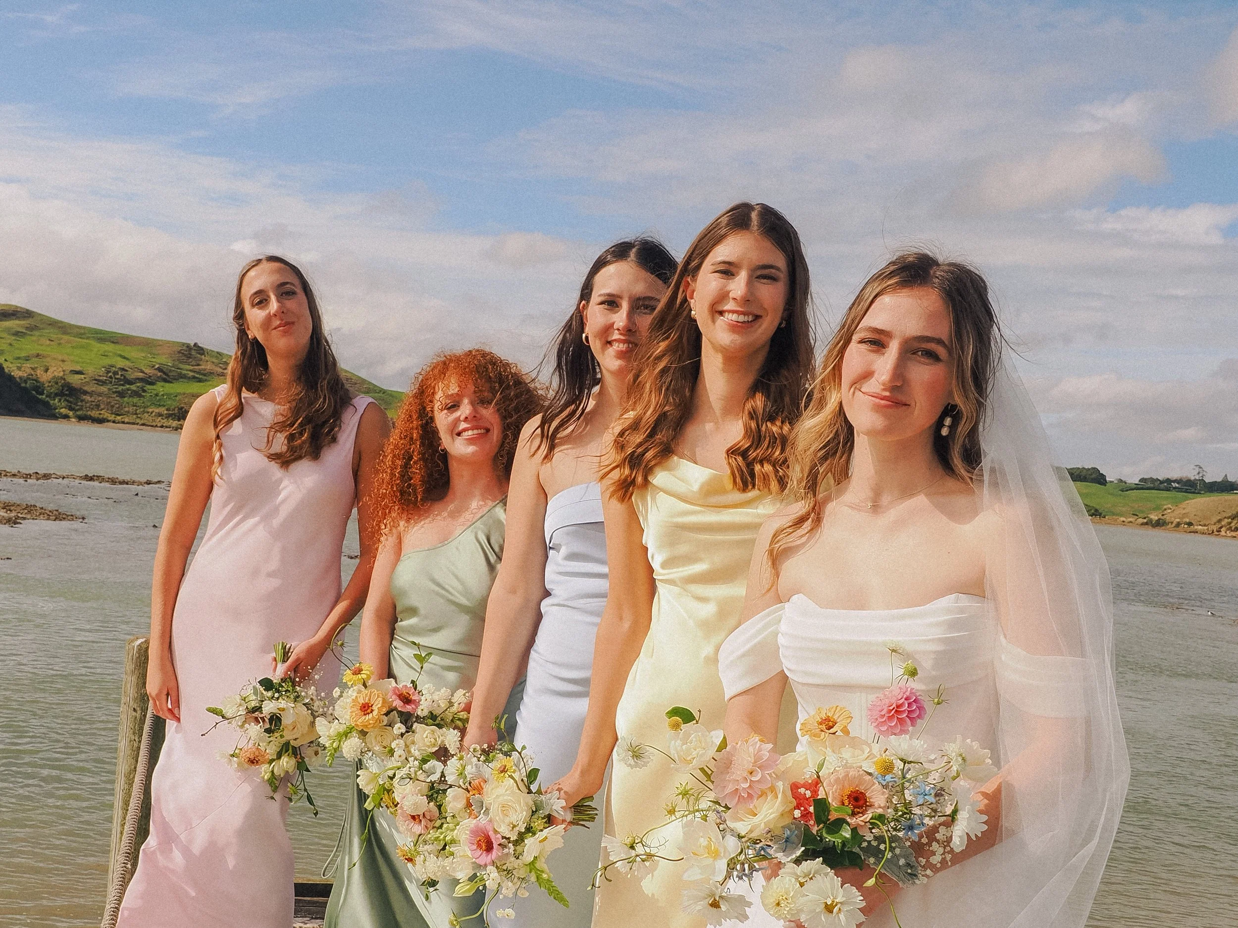 Group of five women in elegant dresses holding bouquets, standing outdoors near water with hills in the background, smiling on a sunny day.  Wedding Photography Waikato New Zealand. Wedding Photography Hamilton New Zealand. Northland Wedding Photo