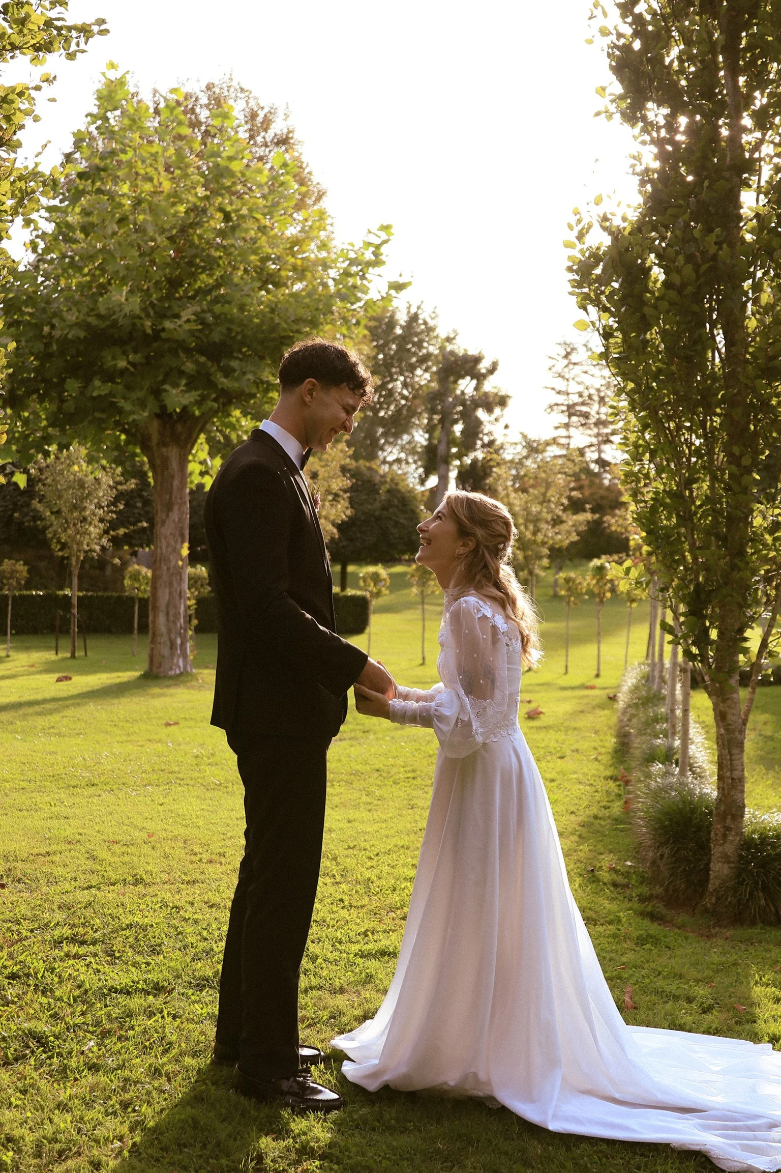 A bride and groom holding hands and smiling at each other in a park with trees and sunlight. Wedding Photography Waikato New Zealand. Wedding Photography Hamilton New Zealand