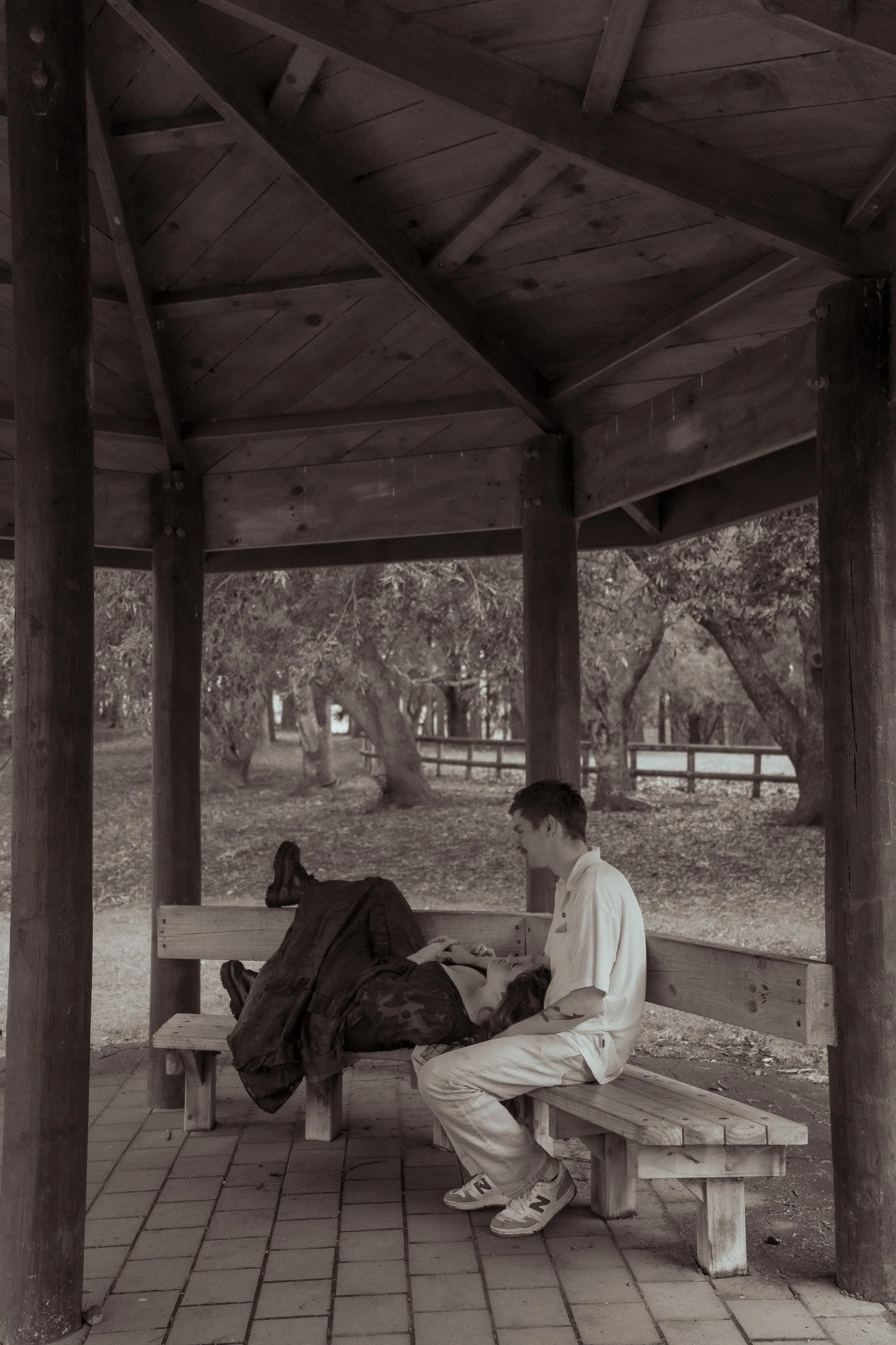 Two teenagers sitting on a wooden park bench under a shelter; the boy is sitting upright and the girl is lying down on the bench with her head on his lap. Couples photography Hamilton New Zealand. Engagement photography Hamilton New Zealand.