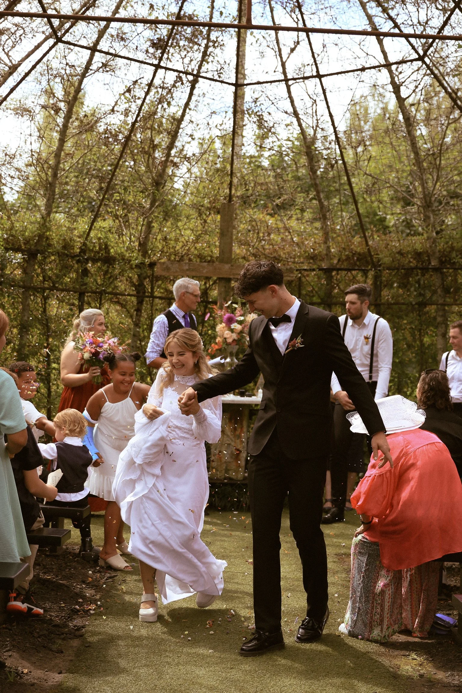 A wedding ceremony taking place outdoors with a bride and groom celebrating with guests, including children and adults, under a rustic open framework surrounded by trees. Wedding Photography Waikato New Zealand. Wedding Photography Hamilton 