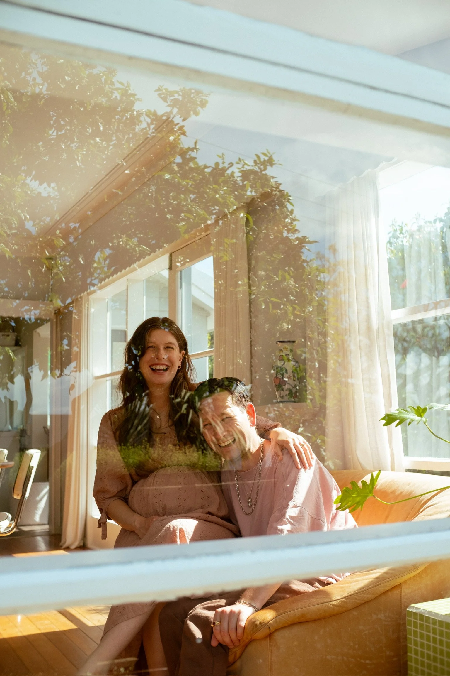 A photo taken through a window showing a laughing woman and man sitting together inside a sunlit room, with greenery and the sky visible outdoors. Maternity Photography. Portrait Photographer. Hamilton New Zealand Photographer