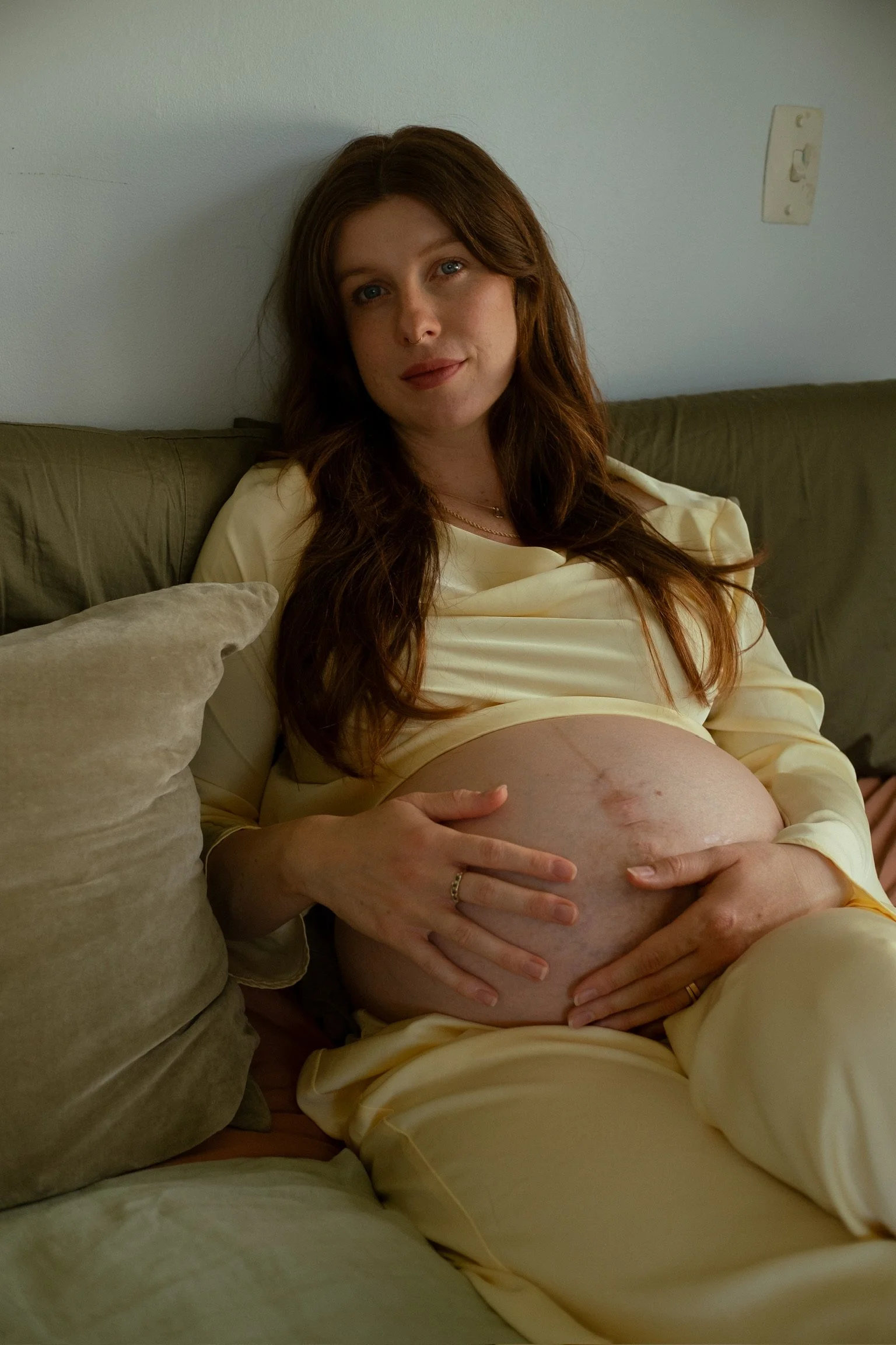 Pregnant woman with long reddish-brown hair sitting on a beige couch, holding her belly and smiling gently, wearing a cream-colored satin dress in a cozy indoor setting. Maternity Photography. Hamilton New Zealand Photographer