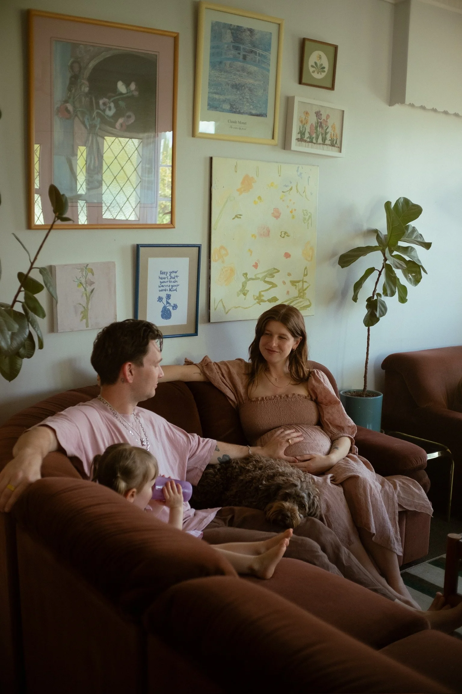 A family sitting on a brown couch in a living room with art on the wall, a pregnant woman, a man, two children, and a dog. Family Photographer. Maternity Photography