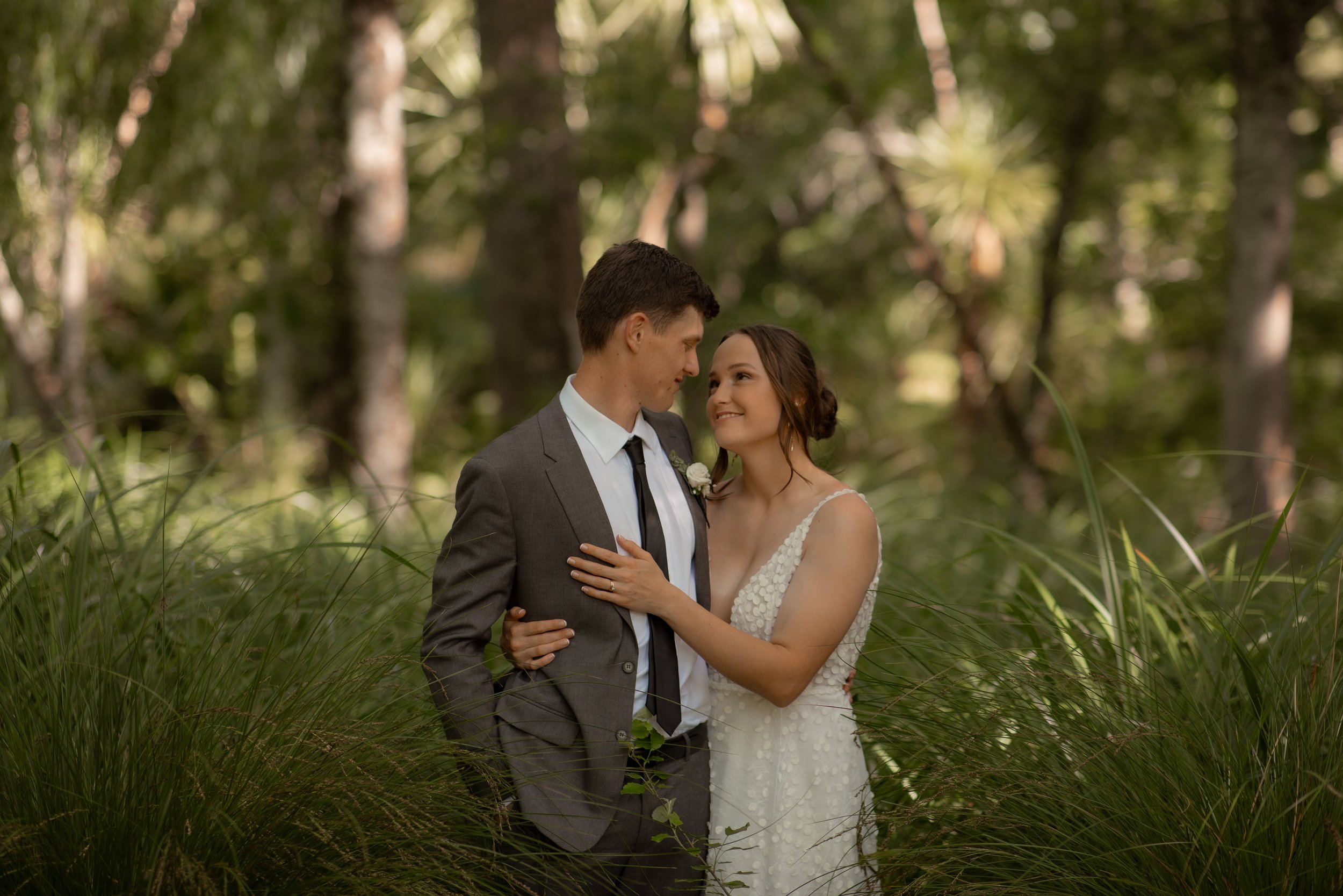 A bride and groom standing close together in a forest, gazing into each other's eyes, surrounded by tall green grass and trees.  Wedding Photography Waikato New Zealand. Wedding Photography Hamilton New Zealand. Northland Wedding Photography