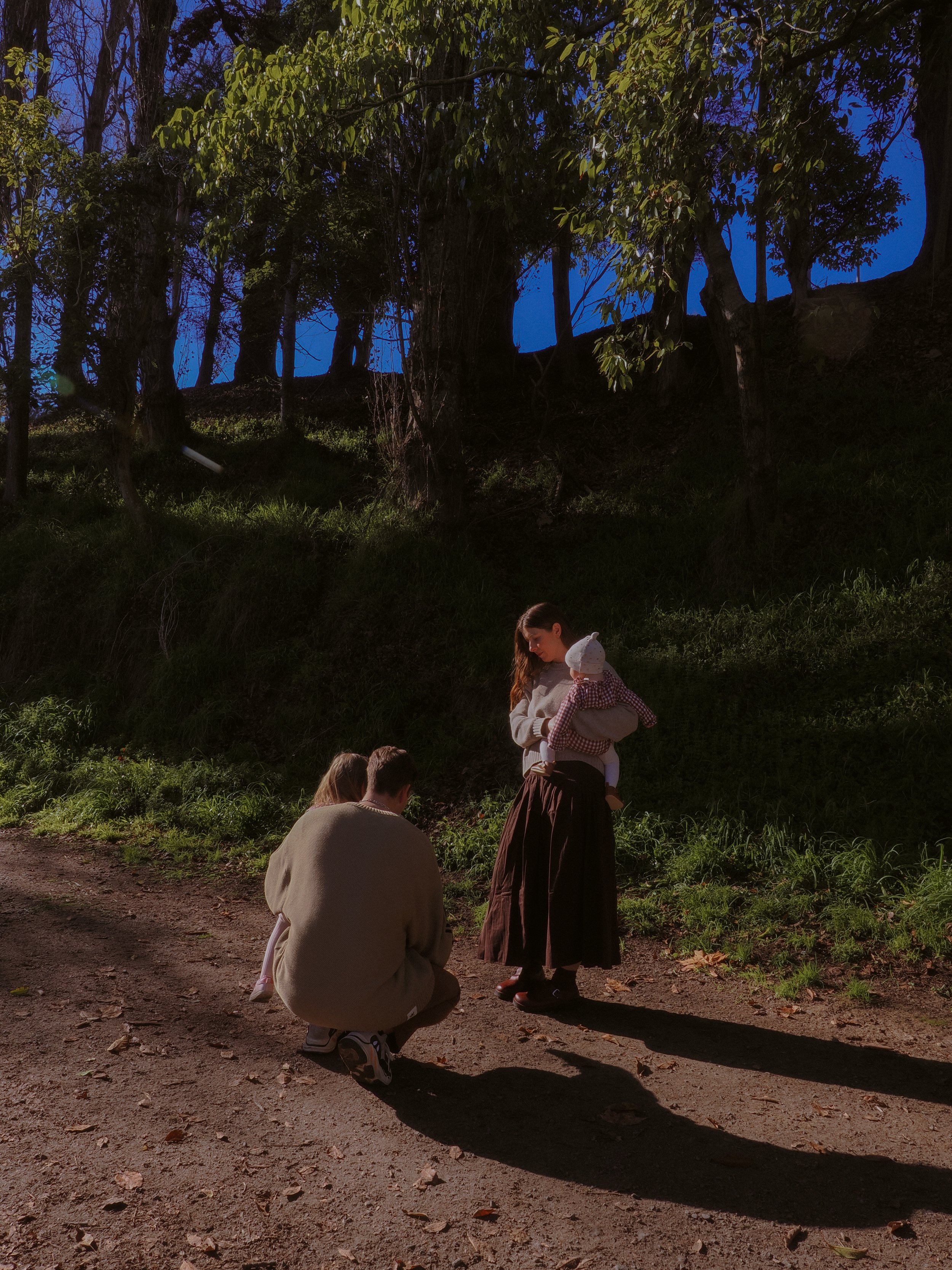 A woman holding a baby standing on a dirt path with two children kneeling in front of her, in a wooded area during twilight. Family Photography Waikato New Zealand. Family Photography Hamilton New Zealand. Northland Wedding Photography