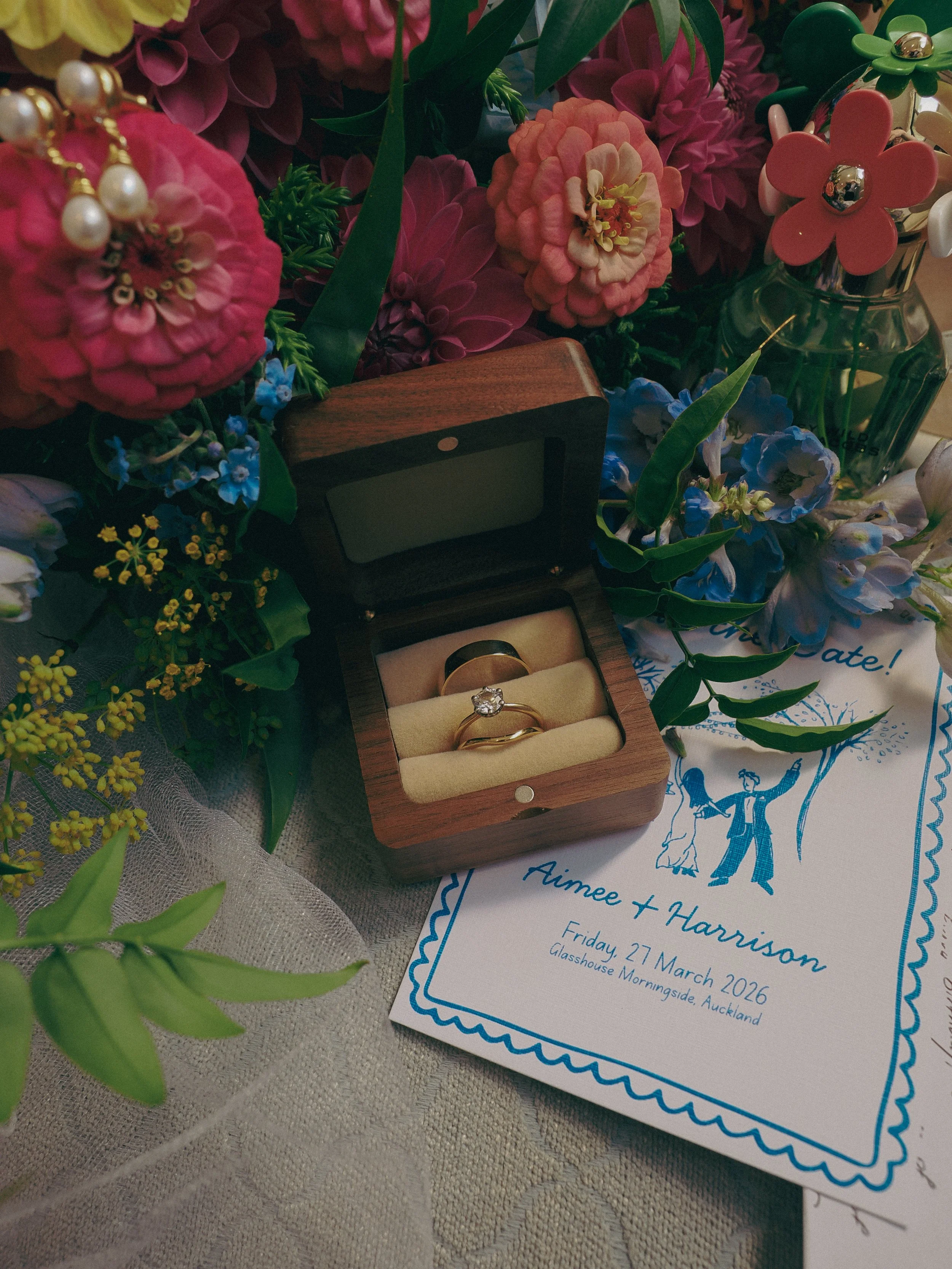 A wedding invitation card for Ameee and Harrison with details of the wedding date and location, surrounded by pink, purple, and blue flowers, a wooden ring box with engagement and wedding rings, and a glass vase with pink and green flowers.