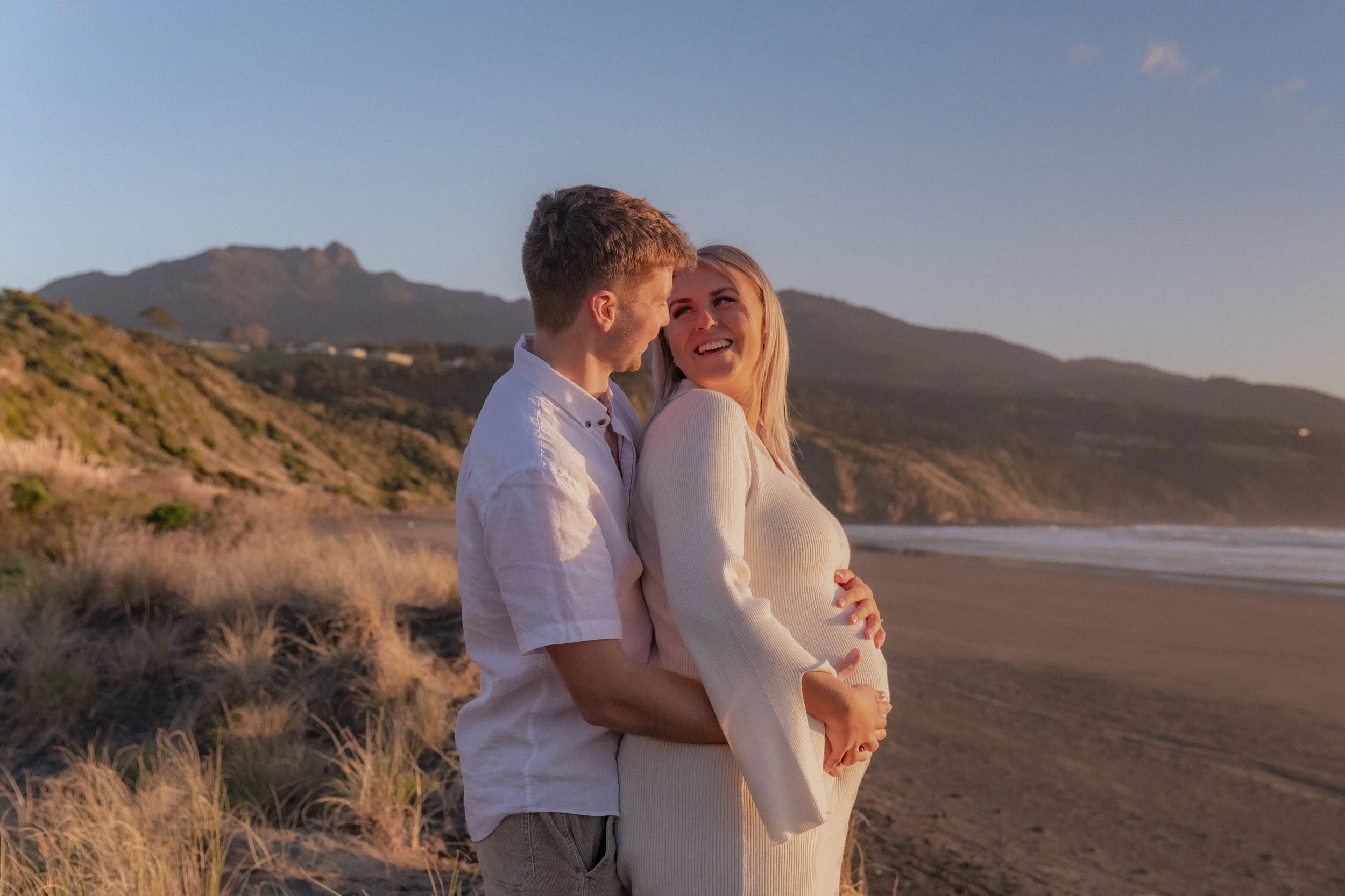 A happy couple, with the man embracing the pregnant woman, standing on a beach during sunset with hills and mountains in the background. Maternity Shoot. Raglan New Zealand