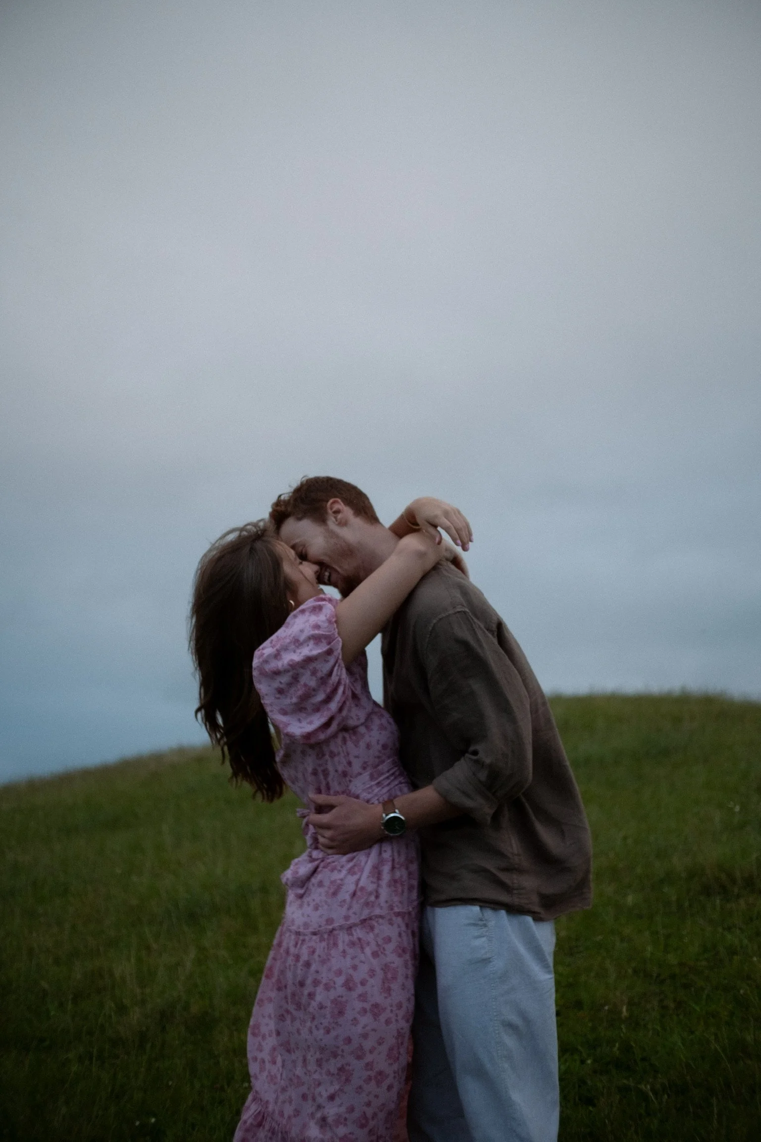 A couple embracing and smiling outdoors on a grassy hill, celebrating a happy moment at dusk. Couples photography Raglan New Zealand. Engagement photography Hamilton New Zealand. Whangarei New Zealand Couples photographer
