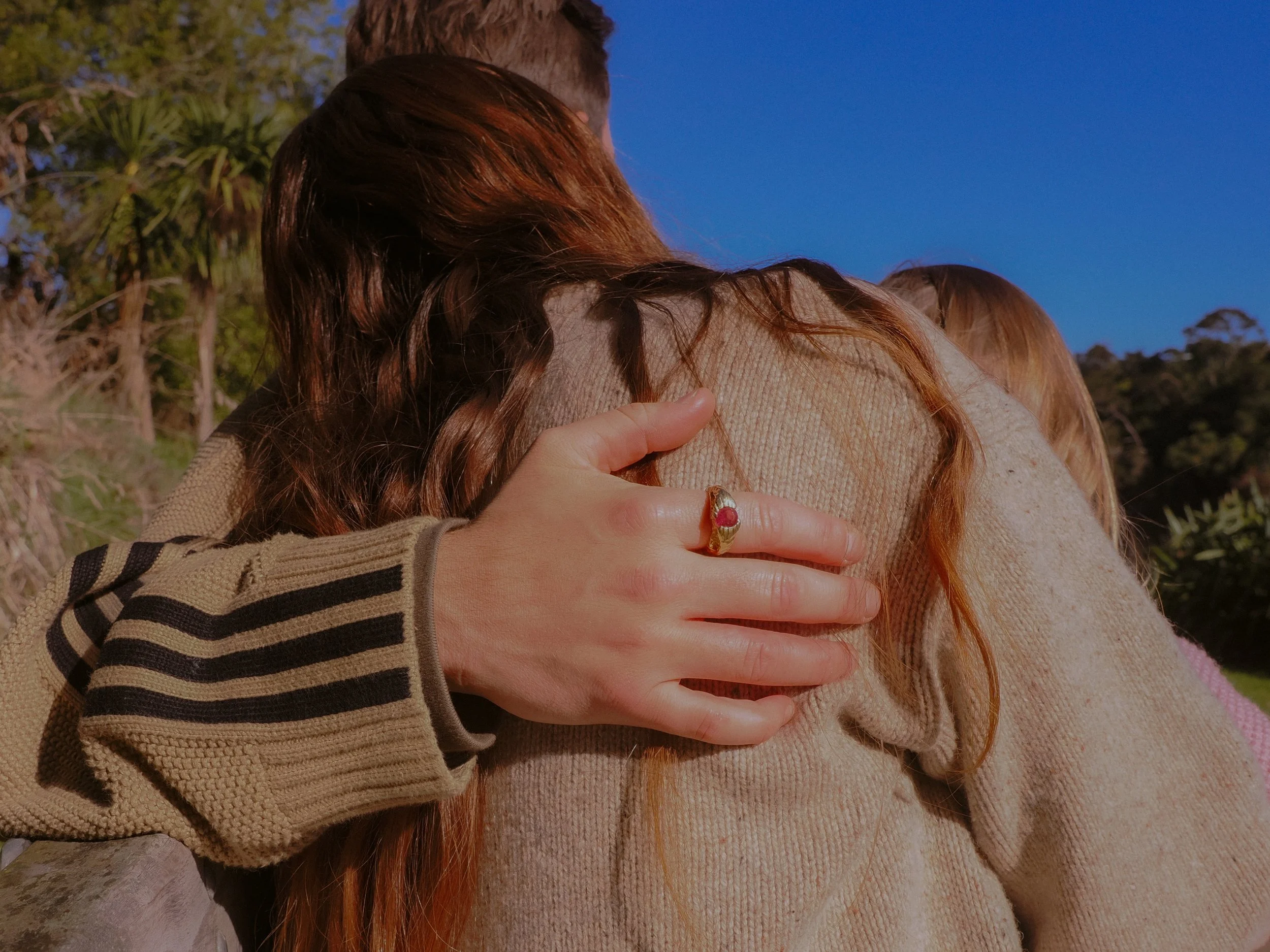 Close-up of two people hugging outdoors, one person's hand resting on the other's shoulder, wearing a ring with a red gemstone, with trees and a blue sky in the background. Family Photography Waikato New Zealand. Family Photography Hamilton