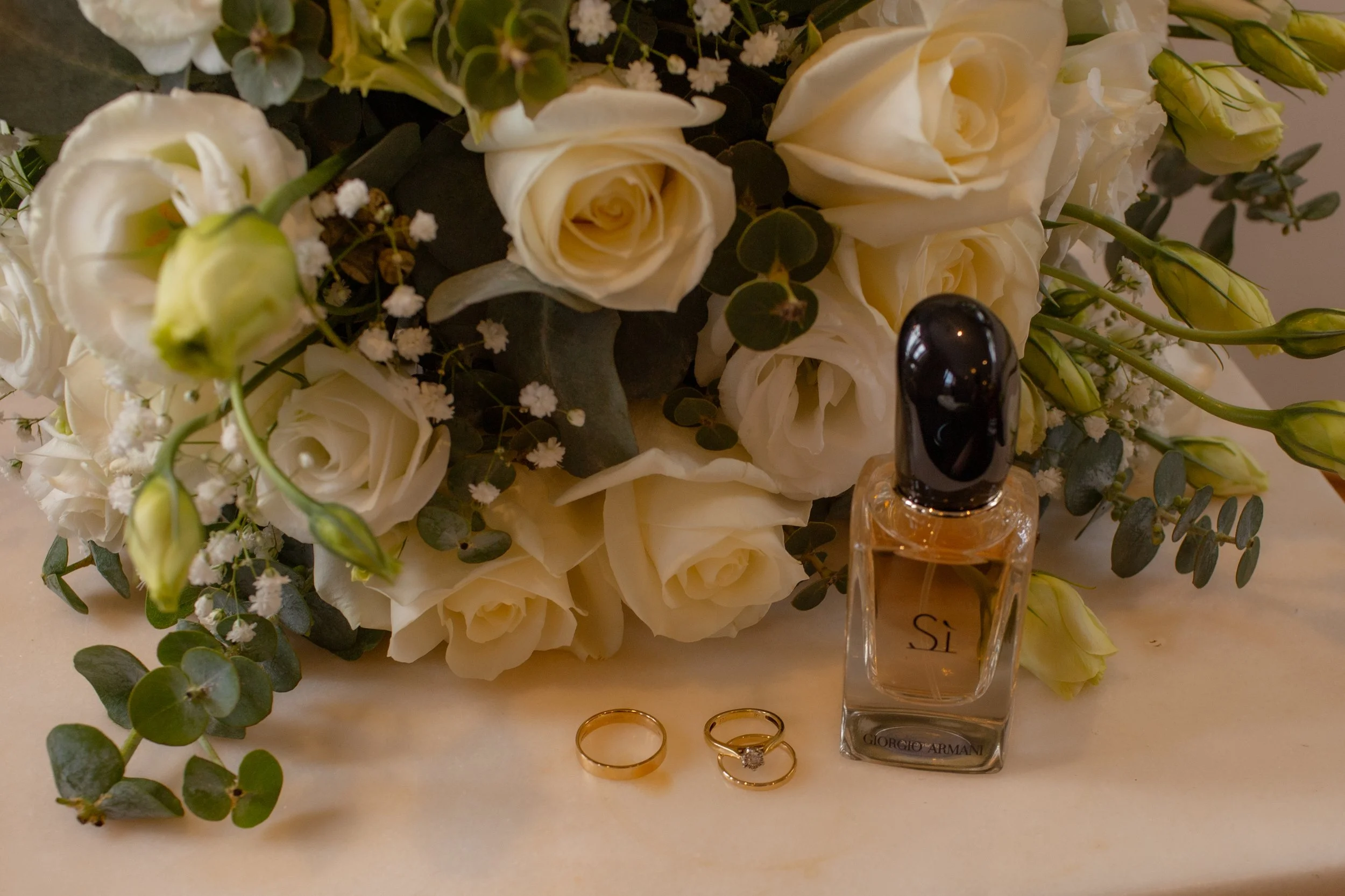 A bouquet of white roses, baby's breath, and eucalyptus leaves next to a perfume bottle and two gold rings on a white surface.  Wedding Photography Waikato New Zealand. Wedding Photography Hamilton New Zealand. Northland Wedding Photography