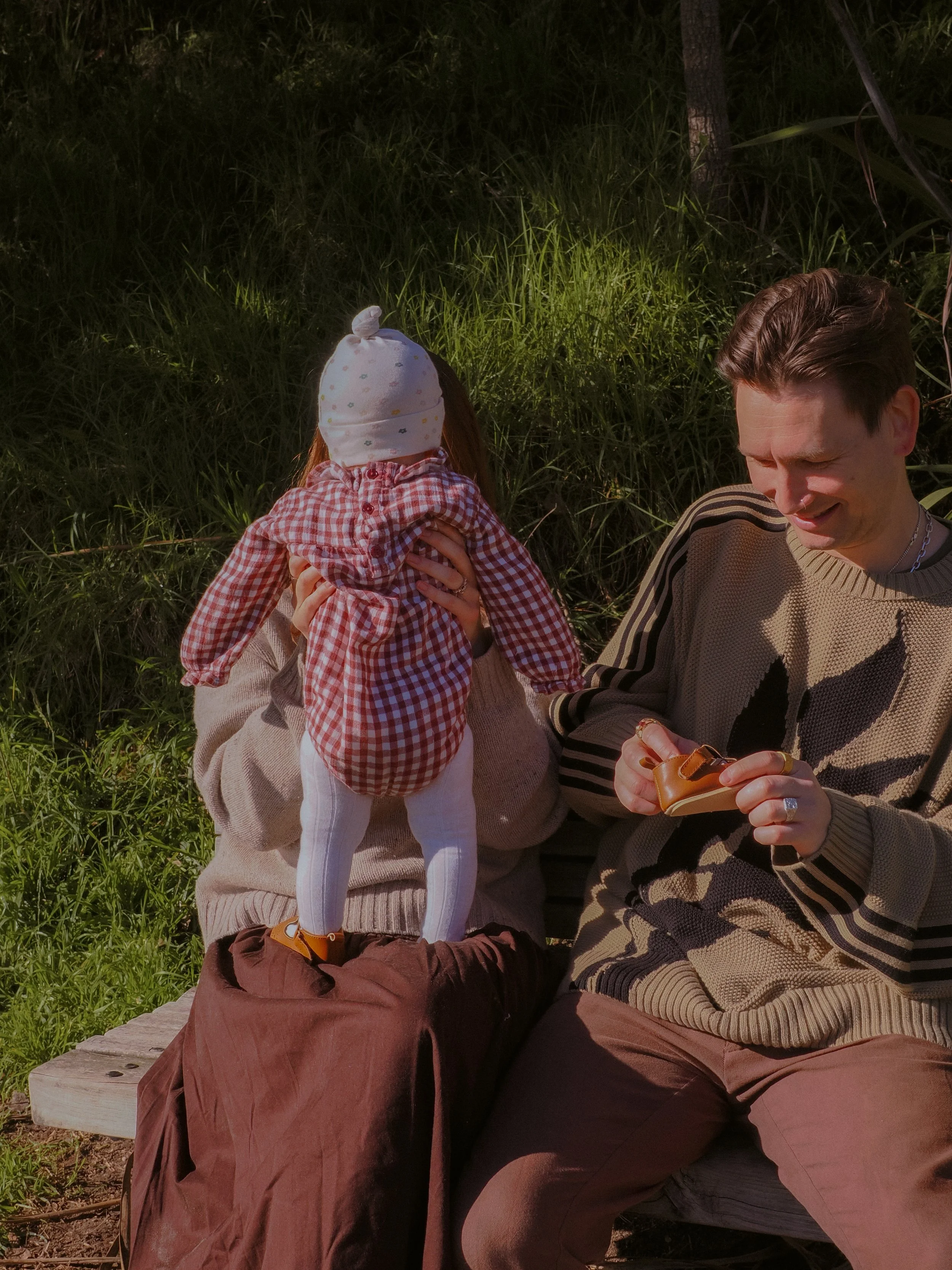 A young girl dressed in a checked jacket, white tights, and a polka dot hat is sitting on an outdoor bench. She is holding a woman, likely her mother, who is sitting beside her. The woman, dressed in a striped sweater, is smiling and looking at a pai