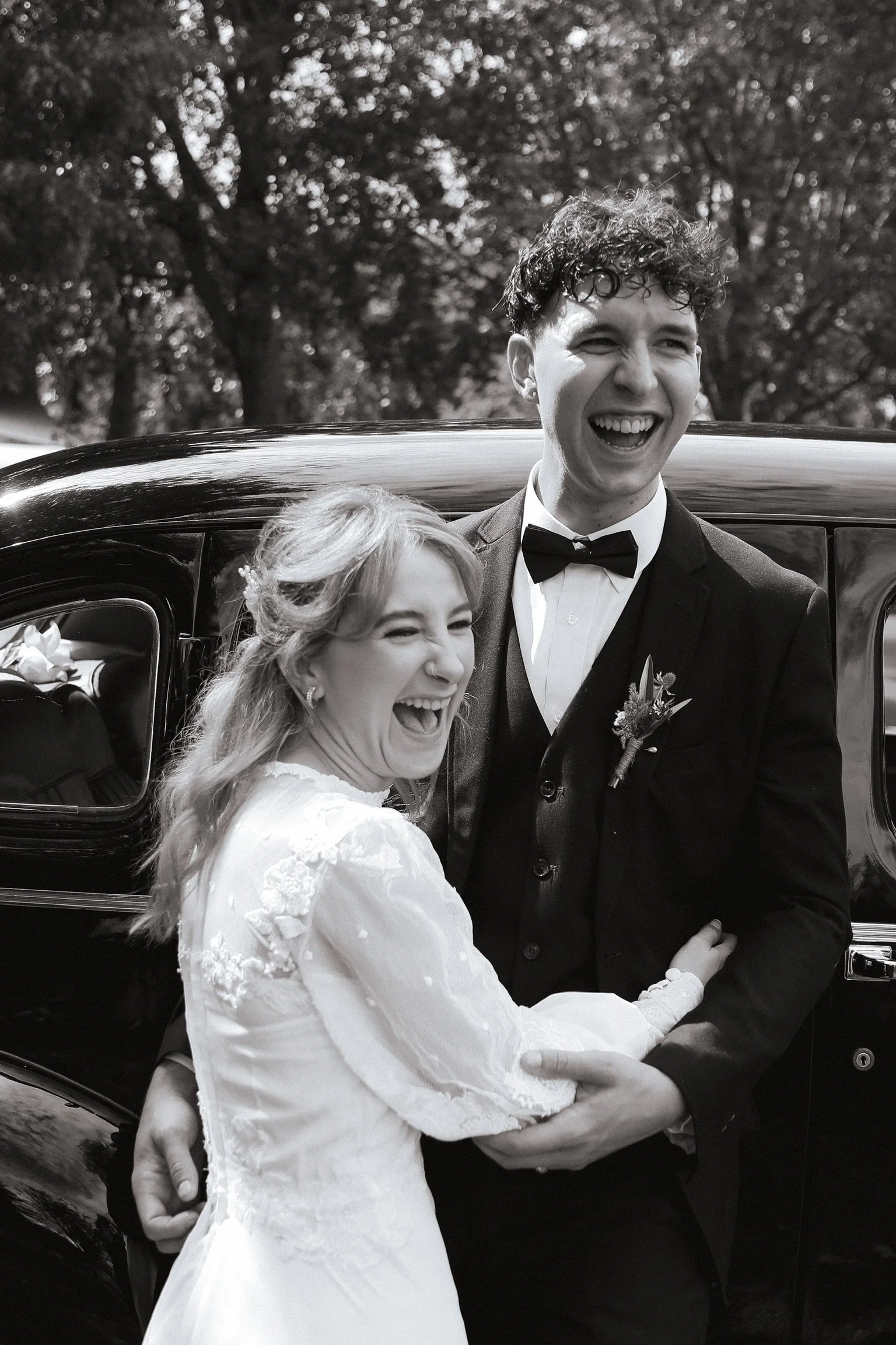 A black and white photo of a happy bride and groom smiling and embracing outdoors, with a vintage black car in the background. Wedding Photography Waikato New Zealand. Wedding Photography Hamilton New Zealand