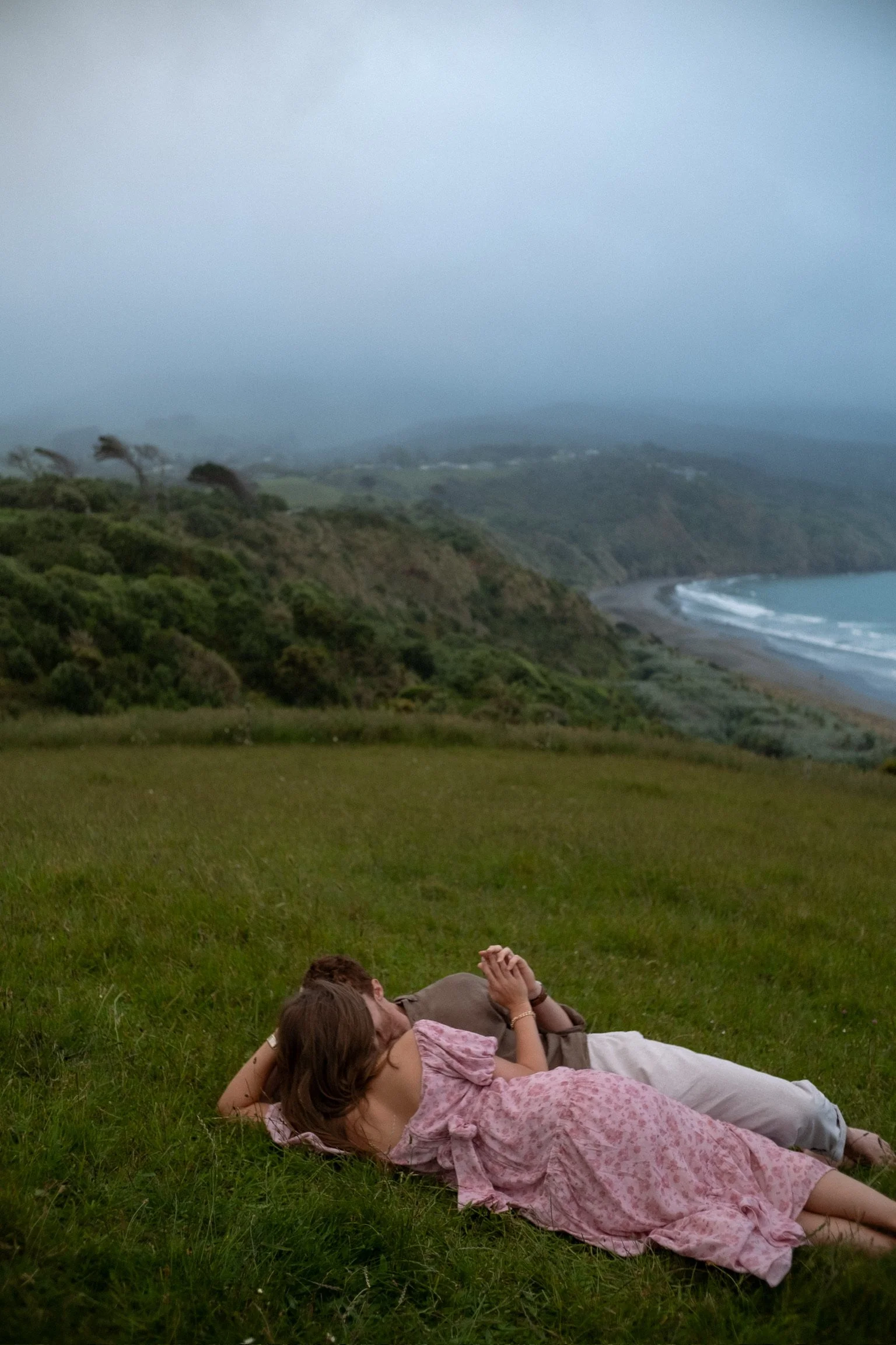 A couple lying on green grass with a coastal landscape, cliffs, and ocean in the background, under a cloudy sky. Couples photography Raglan New Zealand. Engagement photography Hamilton New Zealand. Whangarei New Zealand Couples photographer
