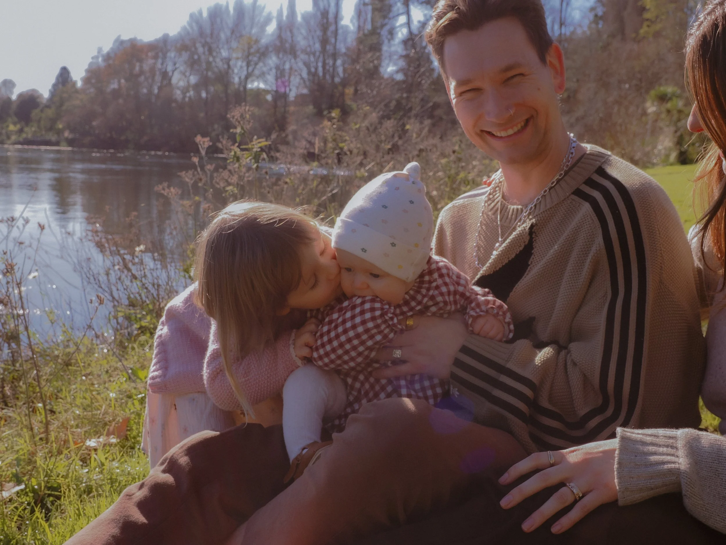A family of four enjoying time outdoors by a lake, with trees in the background. The father is smiling and holding a baby while a young girl kisses the baby's cheek. A woman is partially visible on the right, resting her hand on the father's knee.