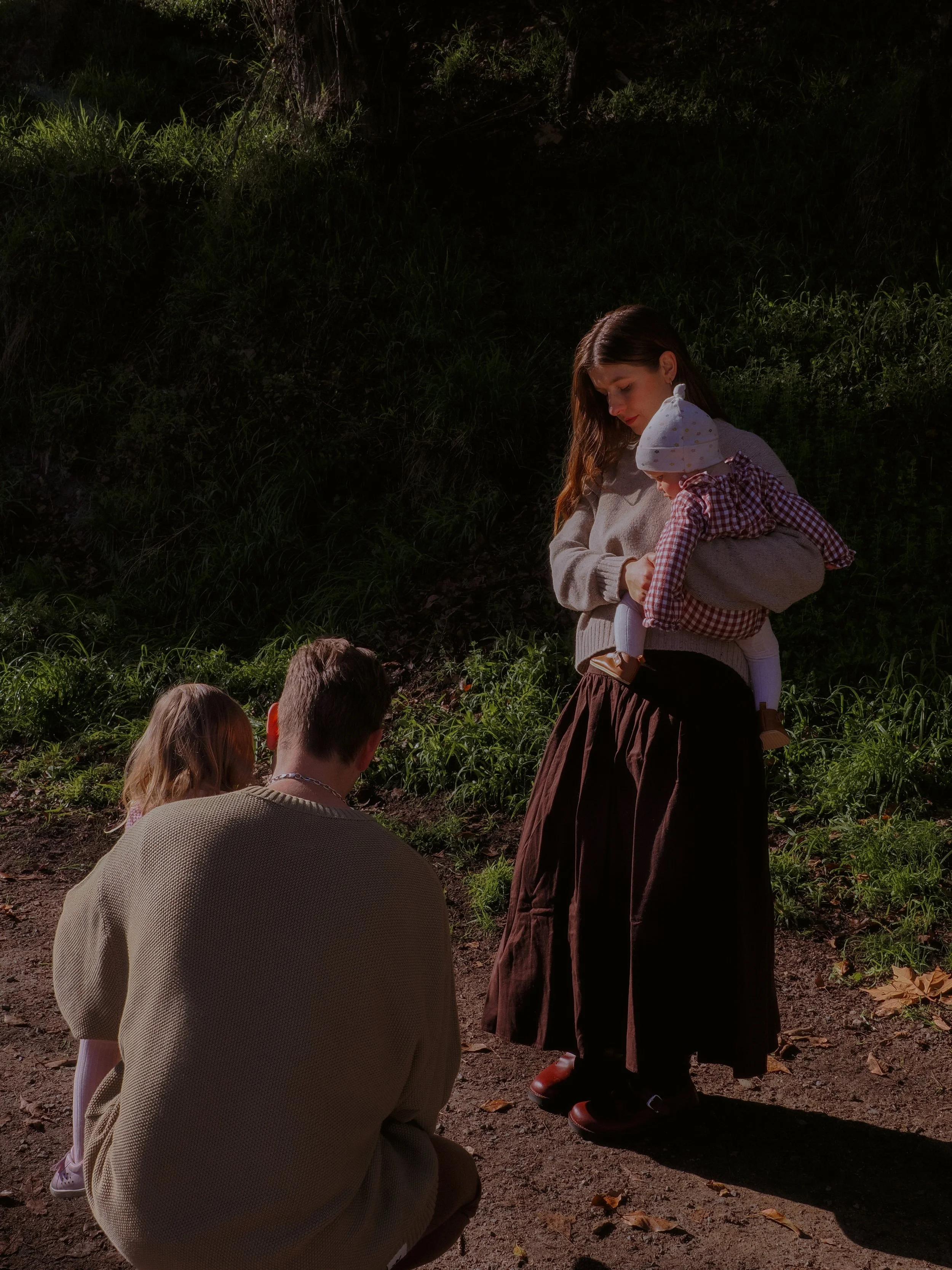 A woman holding a child stands on a dirt path in a forested area, while a man and a girl kneel nearby, engaging in an outdoor activity during daytime. Family Photography Waikato New Zealand. Family Photography Hamilton New Zealand. Northland Wedding 