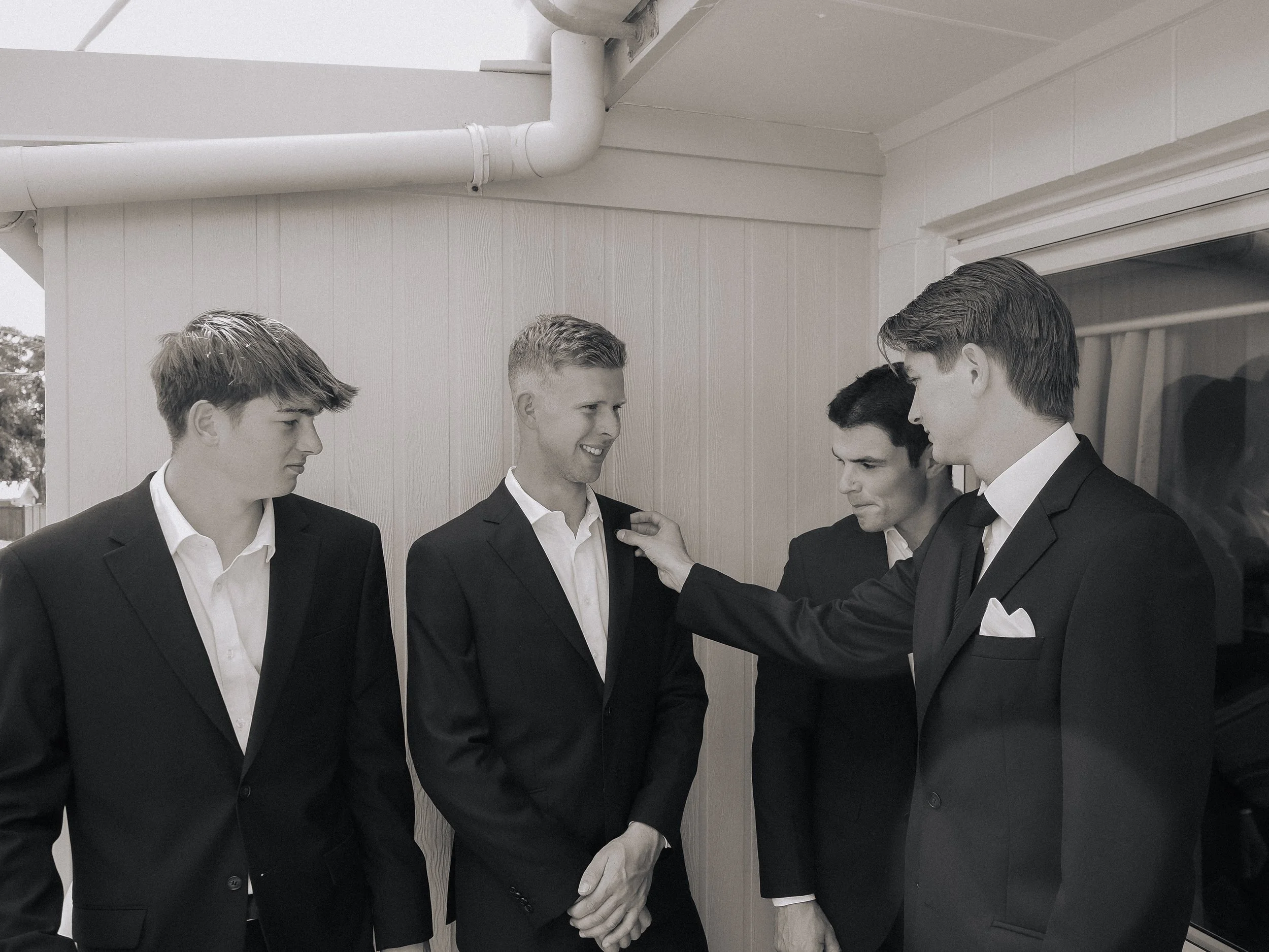 Four men in tuxedos standing close together, engaged in conversation outside near a white building with a window.