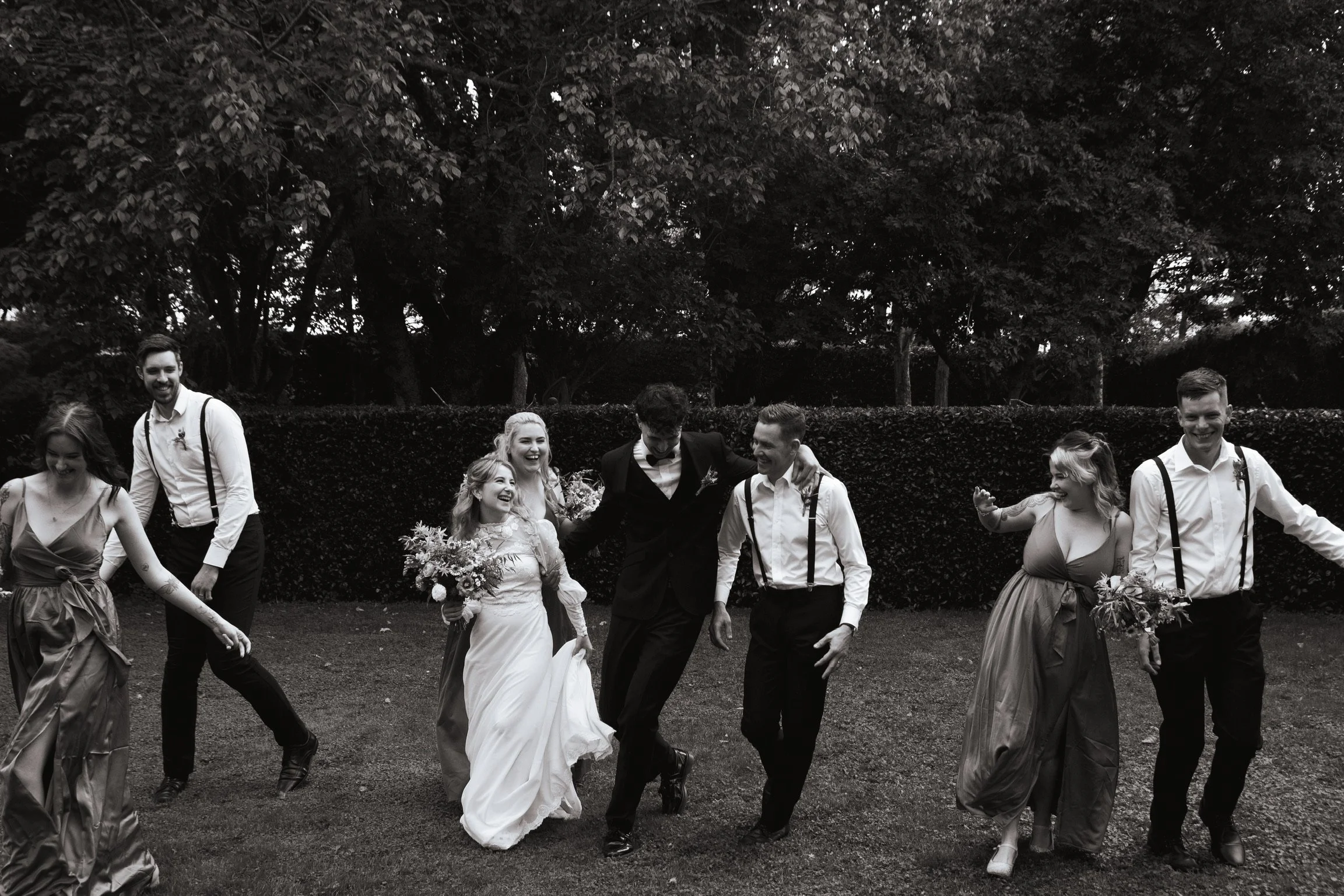 Group of people at a wedding, celebrating and dancing outdoors in a garden with trees and bushes in the background, black and white photograph Wedding Photography Waikato New Zealand. Wedding Photography Hamilton New Zealand