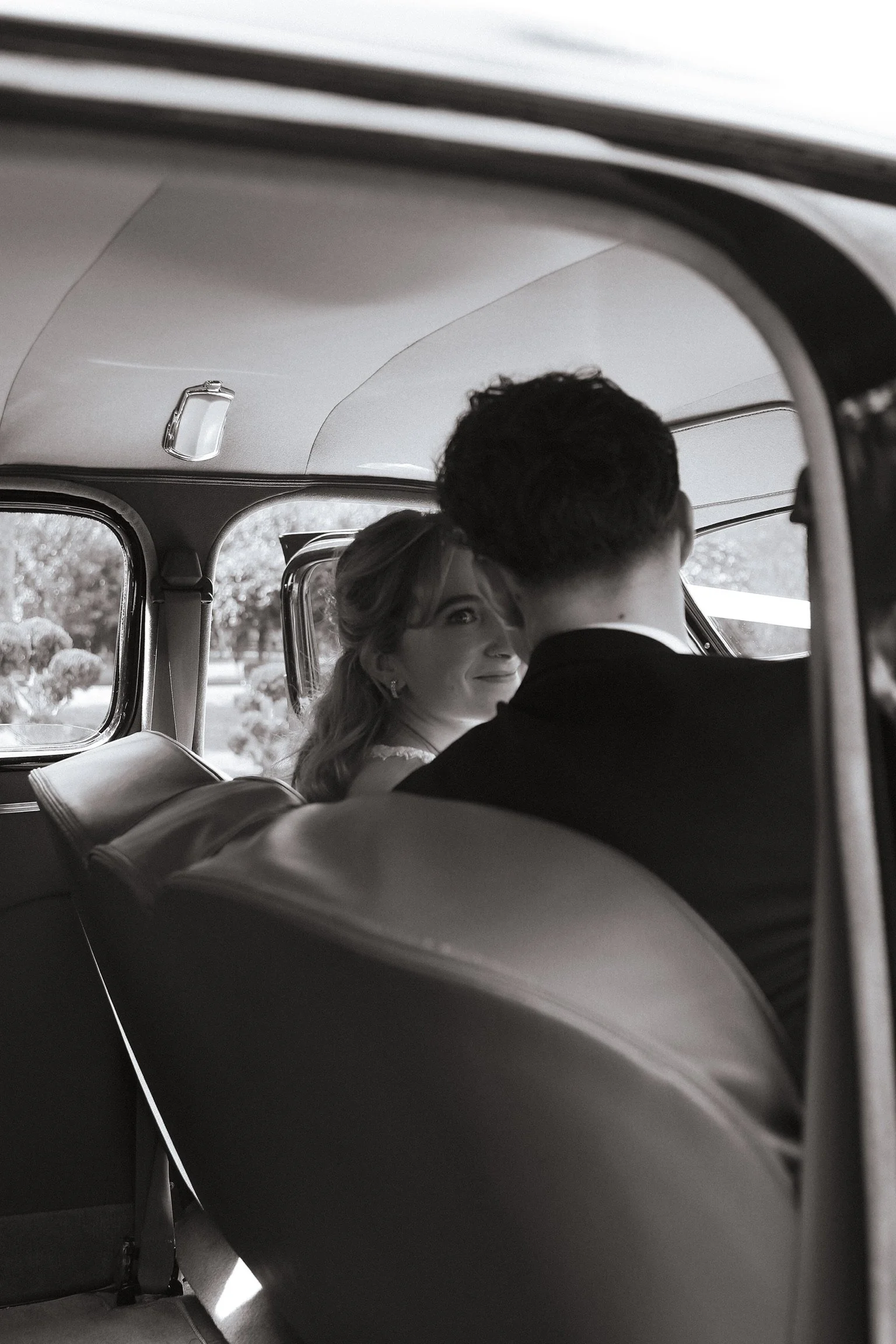 A woman and a man sitting in the backseat of a vintage car, with the woman looking at the man and smiling, captured in black and white. Wedding Photography Waikato New Zealand. Wedding Photography Hamilton New Zealand