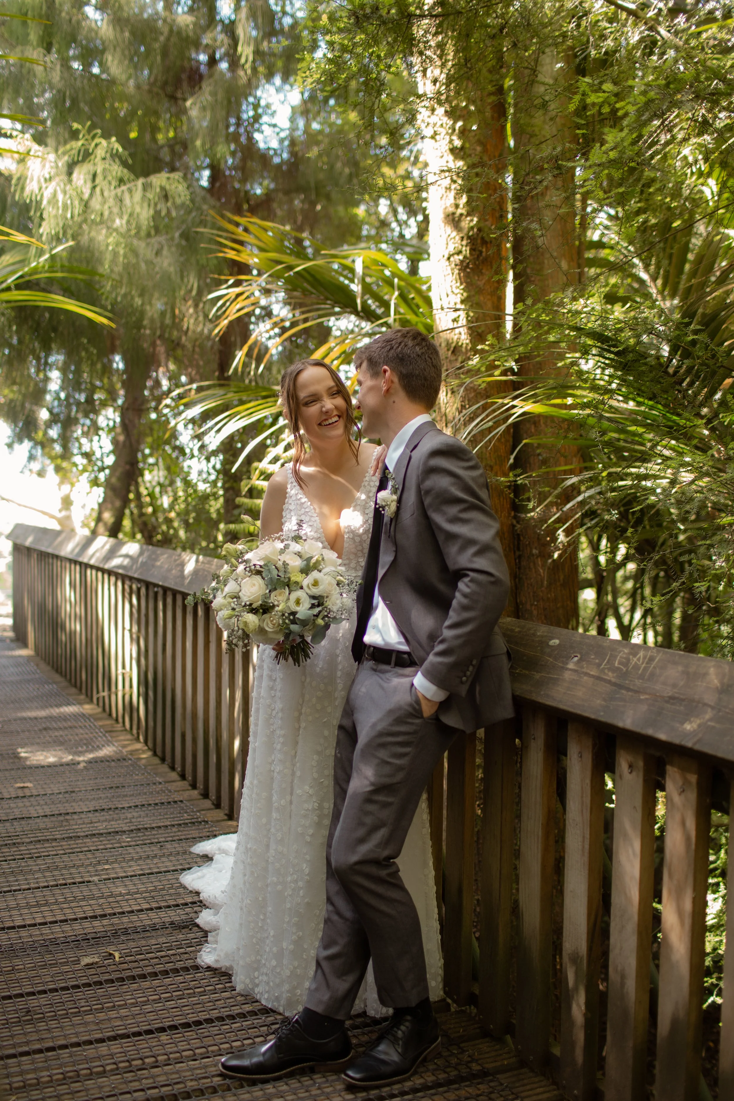 A bride and groom sharing a joyful moment on a wooden bridge surrounded by lush green trees, with the bride holding a bouquet of white flowers.  Wedding Photography Waikato New Zealand. Wedding Photography Hamilton New Zealand. Northland Wedding 