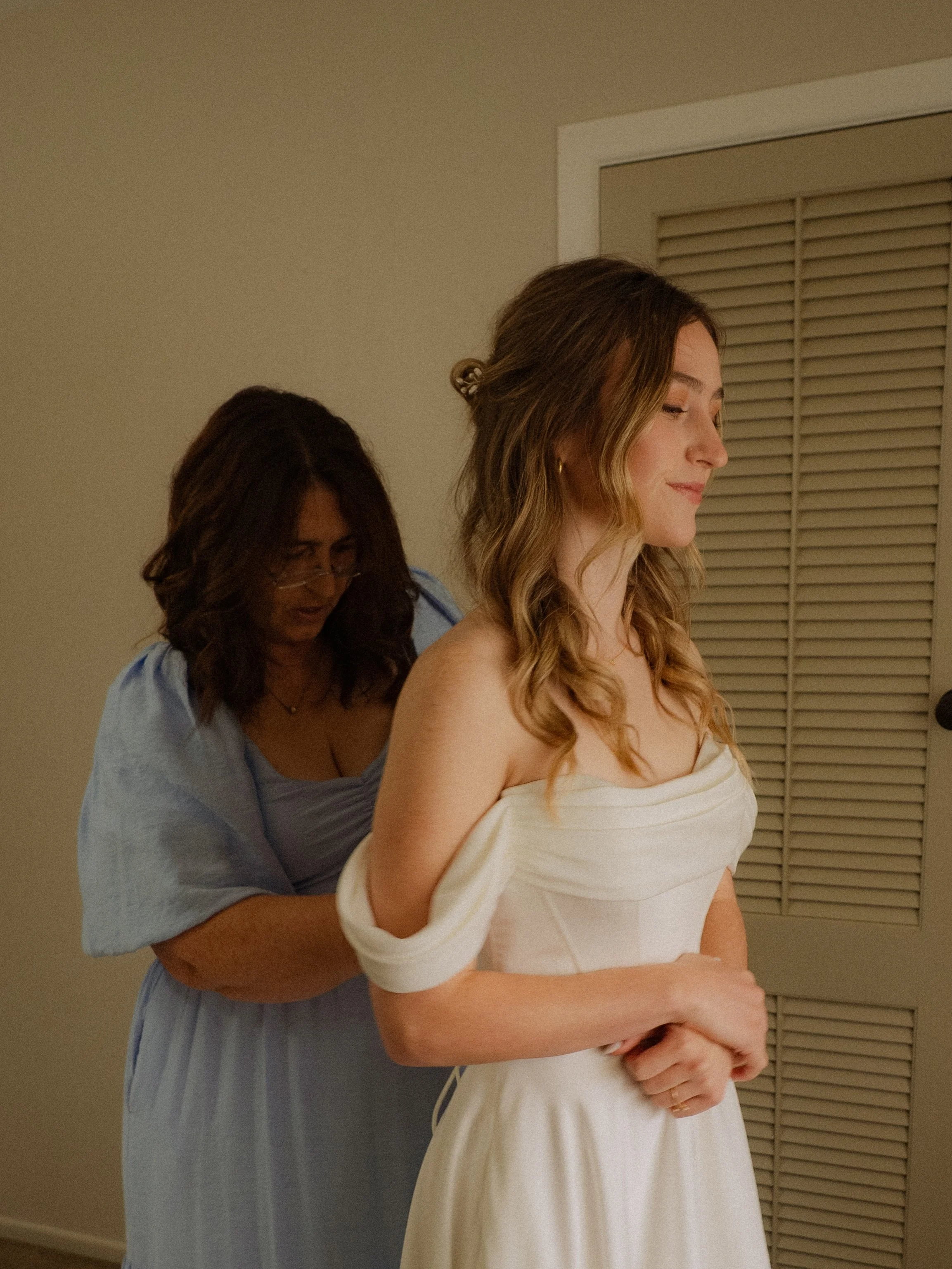 A woman in a white dress is being helped to adjust her dress by another woman in a blue dress.  Wedding Photography Waikato New Zealand. Wedding Photography Hamilton New Zealand. Northland Wedding Photography
