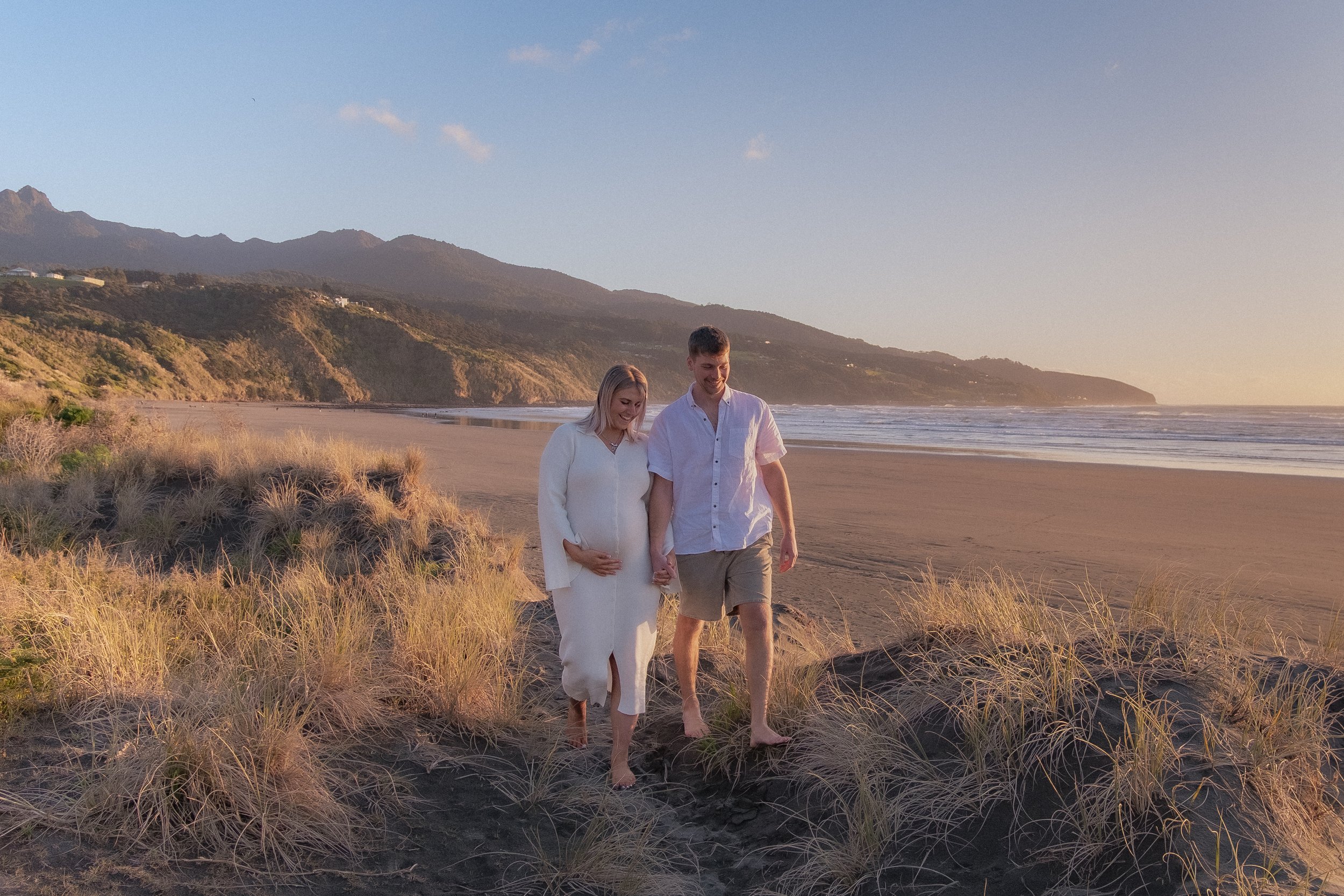 A couple walking barefoot on a sandy beach with sand dunes and beach grass, holding hands, during sunset with mountains in the background. Maternity Shoot. Raglan New Zealand