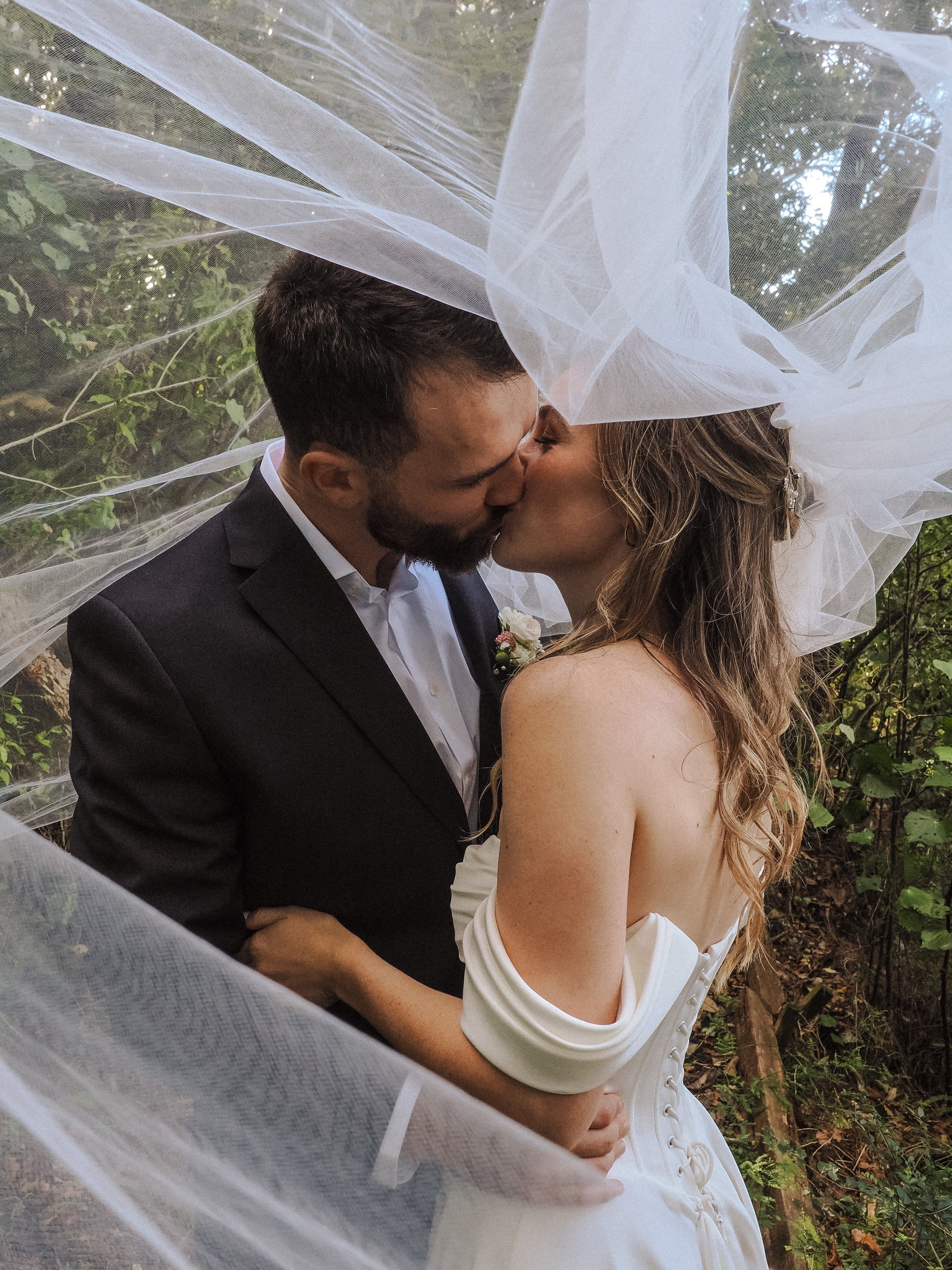 A couple in wedding attire sharing a kiss outdoors, surrounded by a sheer white veil and green foliage.