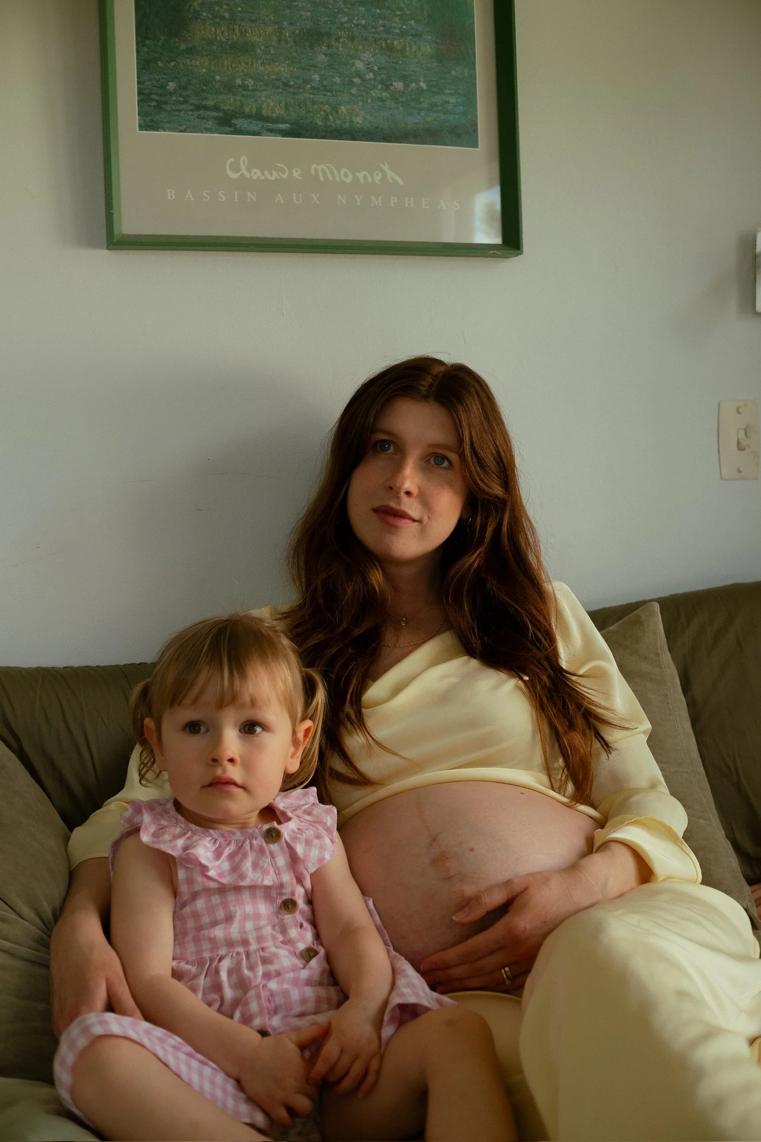 A pregnant woman with long brown hair sitting on a couch, holding her belly, with a young girl with blonde hair and a pink dress sitting next to her. Maternity Photography. Hamilton New Zealand Photographer