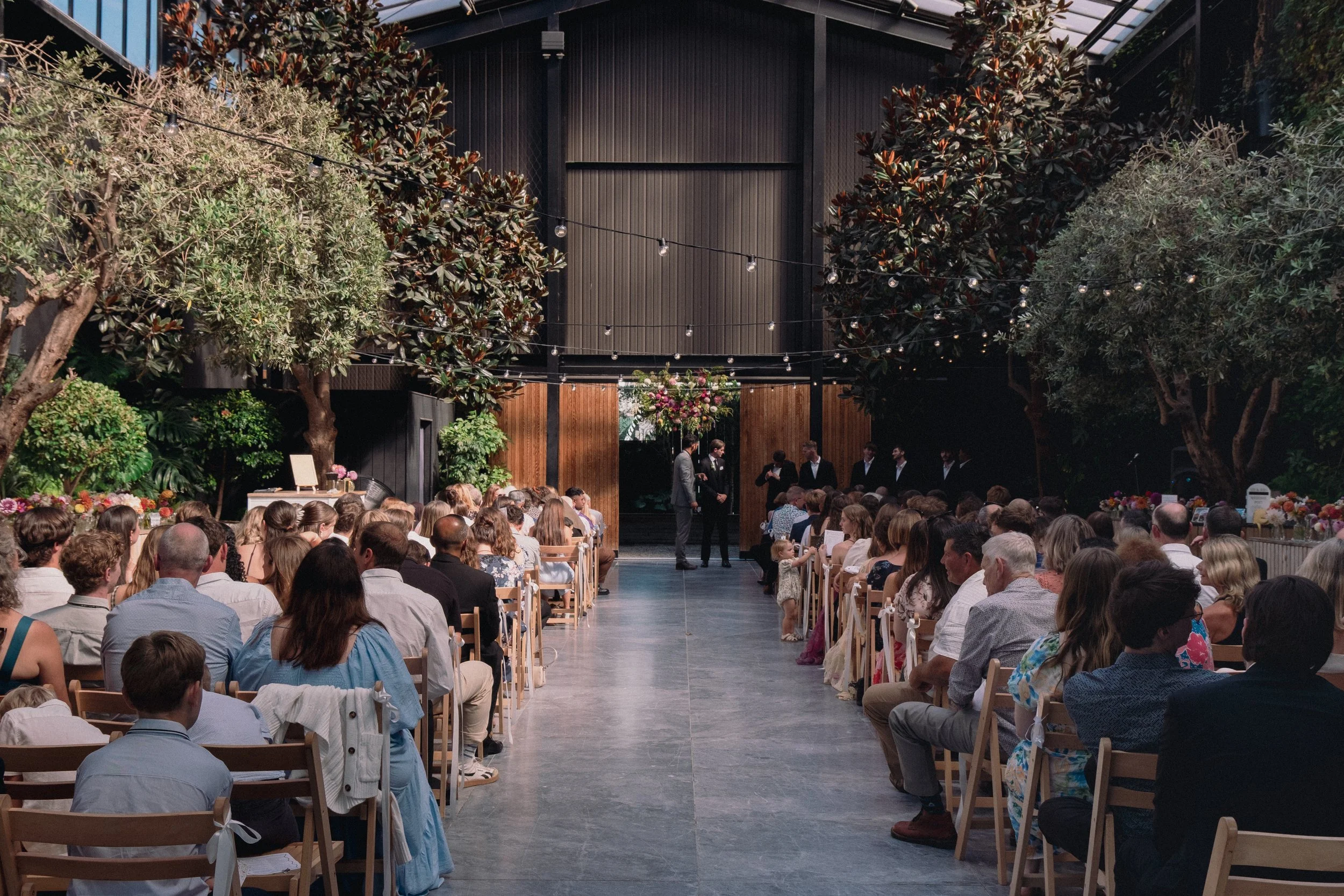 A wedding ceremony inside a large, modern, industrial-style venue with a high black ceiling, wooden wall panels, and string lights. Guests are seated on wooden chairs, facing a small stage where a couple and officiant are standing, with the bride and