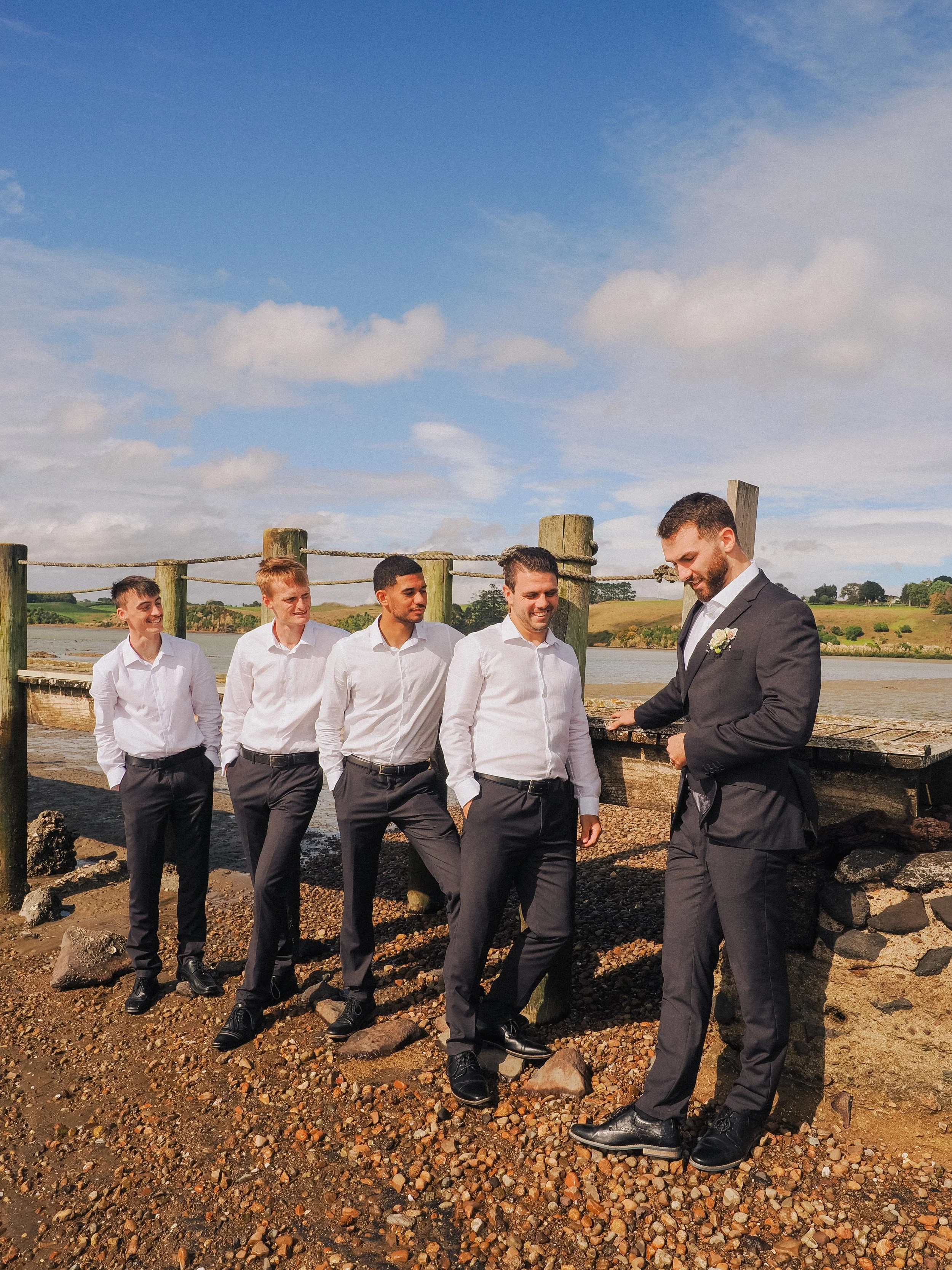 A group of five men in formal attire standing outdoors near a body of water, with a blue sky and scattered clouds in the background. The man on the right, dressed in a suit, is looking down and smiling, while the others are smiling and looking at him