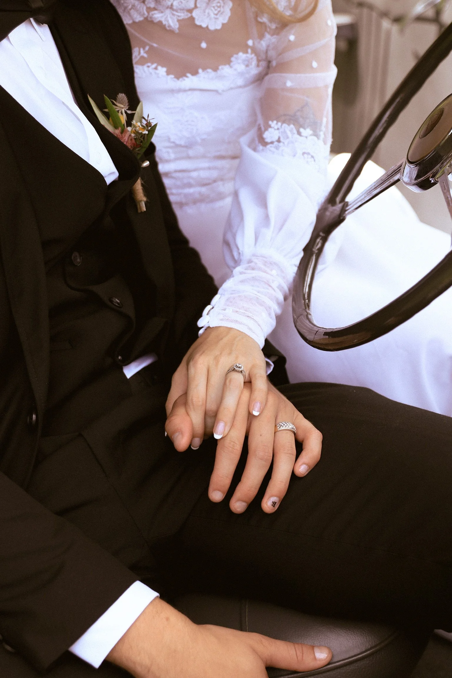 Close-up of a bride and groom holding hands, showing wedding rings, with the bride wearing lace sleeves and the groom in a dark suit with a boutonnière. Wedding Photography Waikato New Zealand. Wedding Photography Hamilton New Zealand