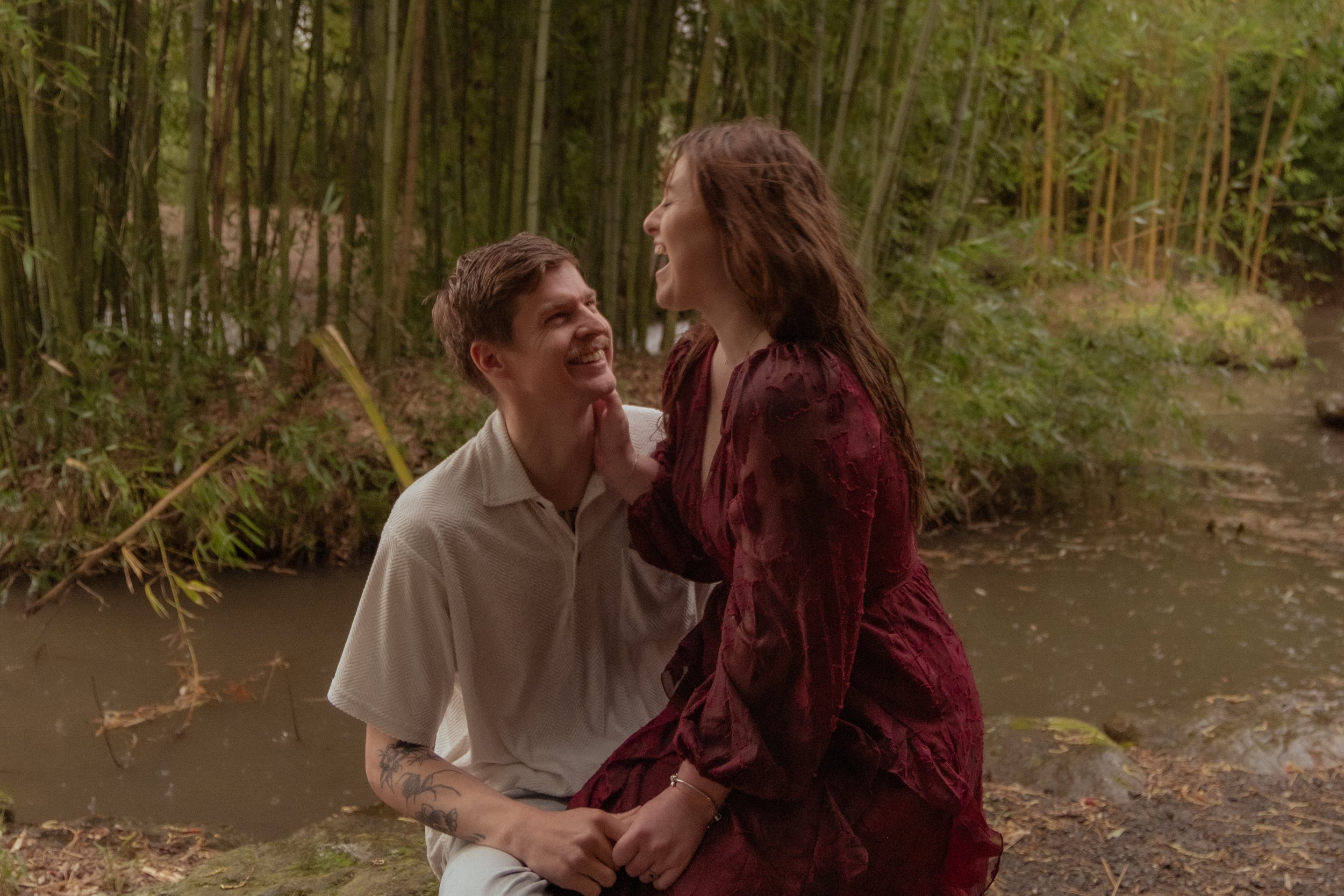A couple sitting by a creek in a bamboo forest, smiling and looking at each other. The woman is touching the man's chin and has long brown hair and a red dress. The man has short brown hair, a mustache, and tattoos on his arm.