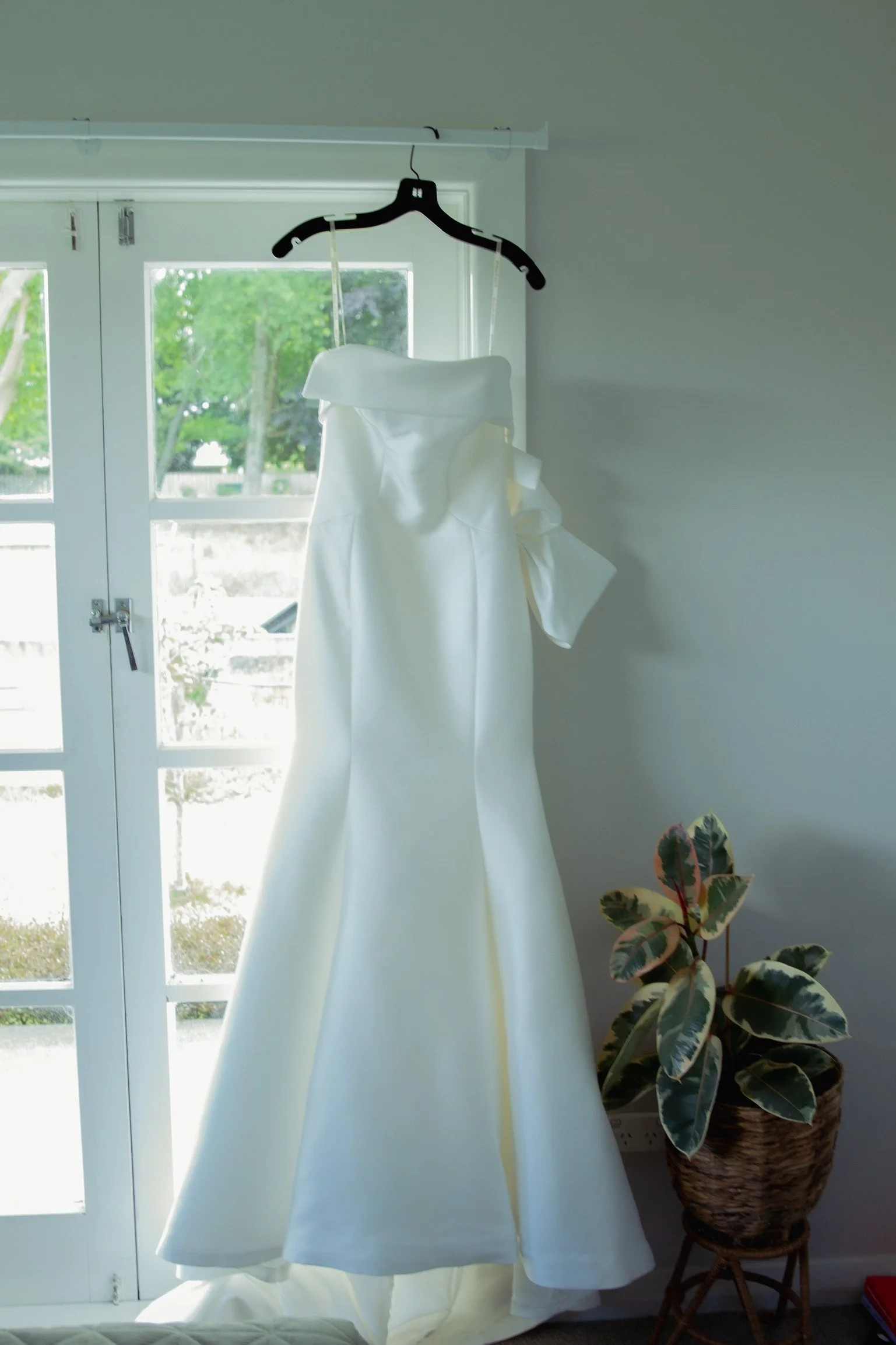 White wedding dress hanging on a black hanger in front of a glass door with greenery outside, next to a potted plant with variegated leaves  Wedding Photography Waikato New Zealand. Wedding Photography Hamilton New Zealand. Northland Wedding Photo