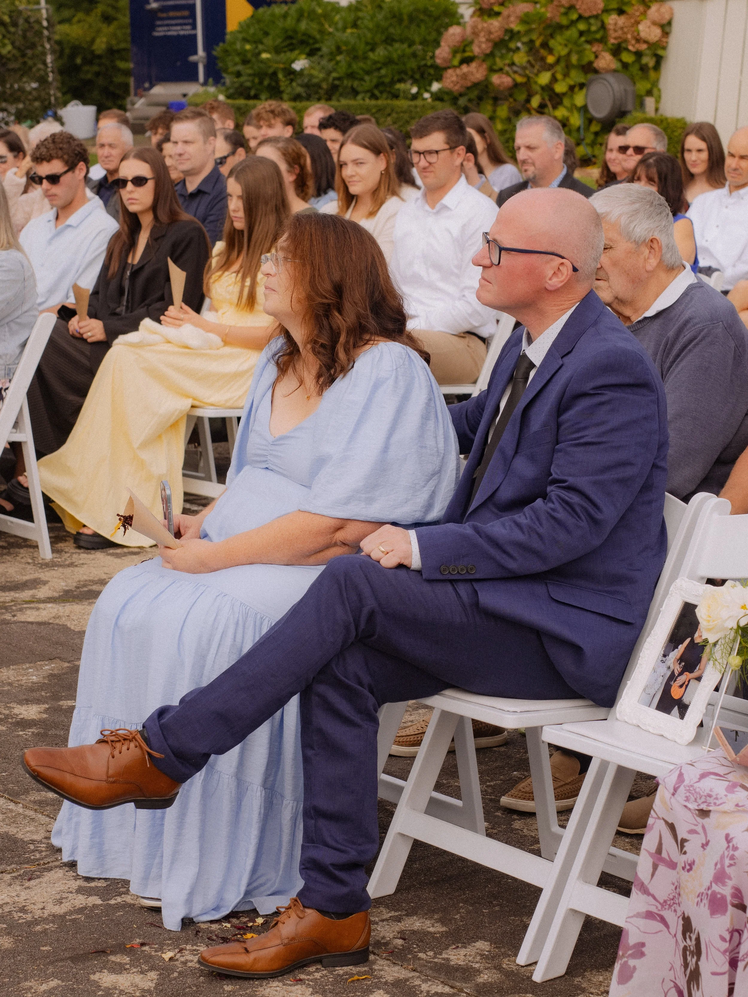 A diverse group of people attending an outdoor event, sitting on white chairs, with some holding programs or photographs, and a floral arrangement and framed photo on a chair in the foreground.  Wedding Photography Waikato New Zealand. 