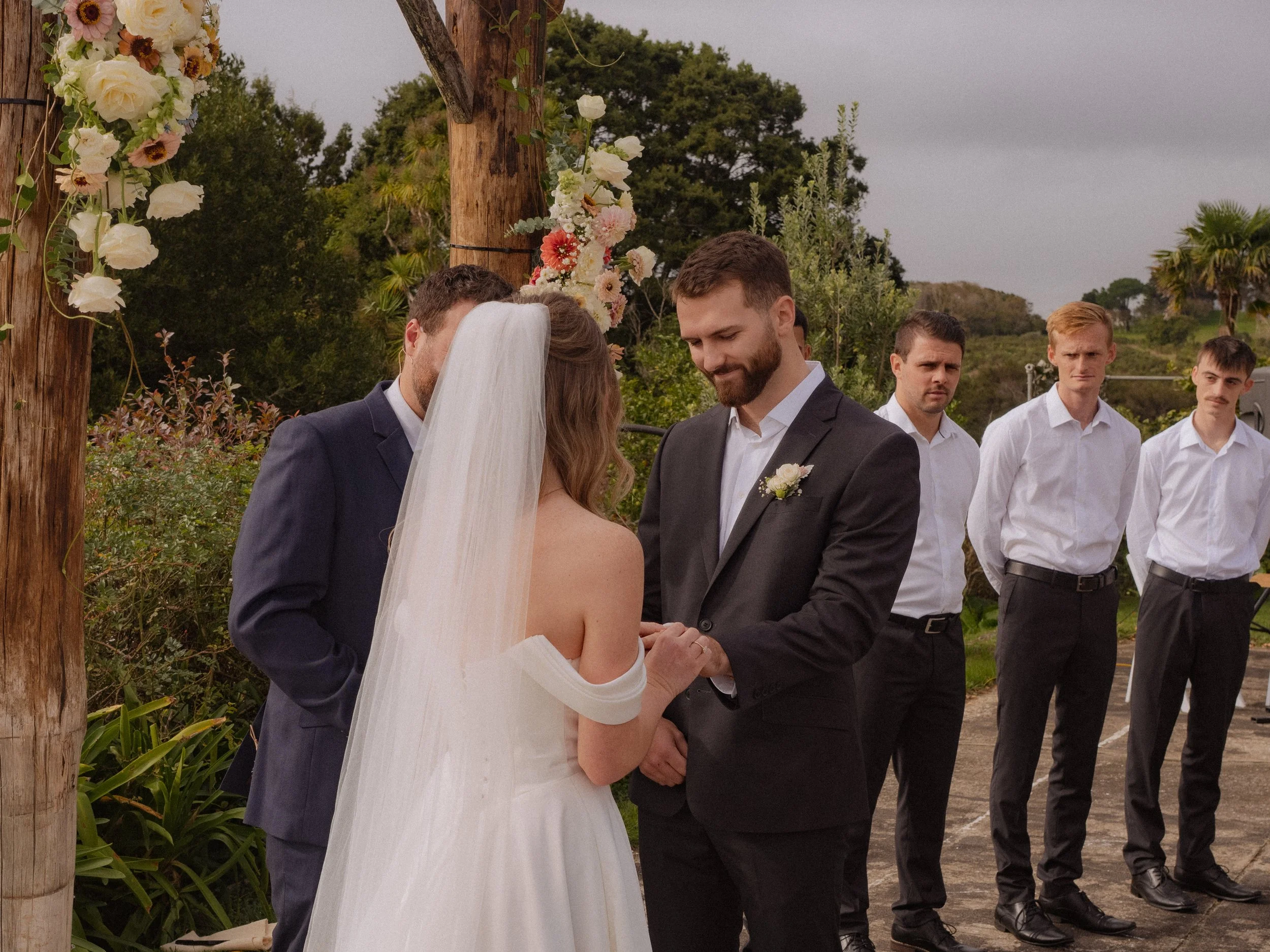 A wedding ceremony outdoors with a bride and groom holding hands, standing before an officiant. The bride wears a white off-shoulder gown and veil, and the groom wears a dark suit with a white shirt and boutonniere. In the background, four groomsmen 