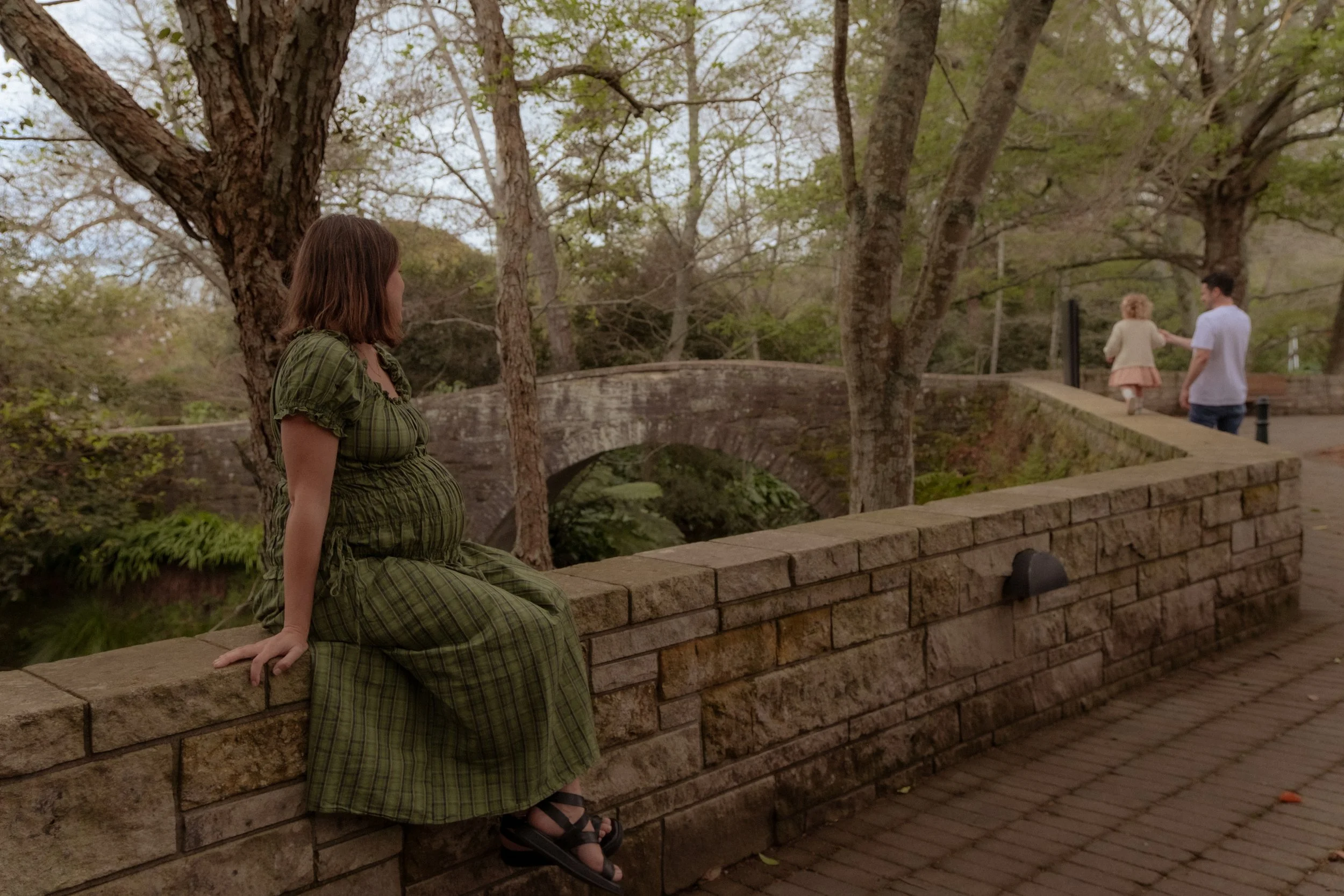 A pregnant woman in a green plaid dress sitting on a stone wall near a bridge in a park, with two children and a man walking away in the background. Family/Maternity photography Hamilton New Zealand