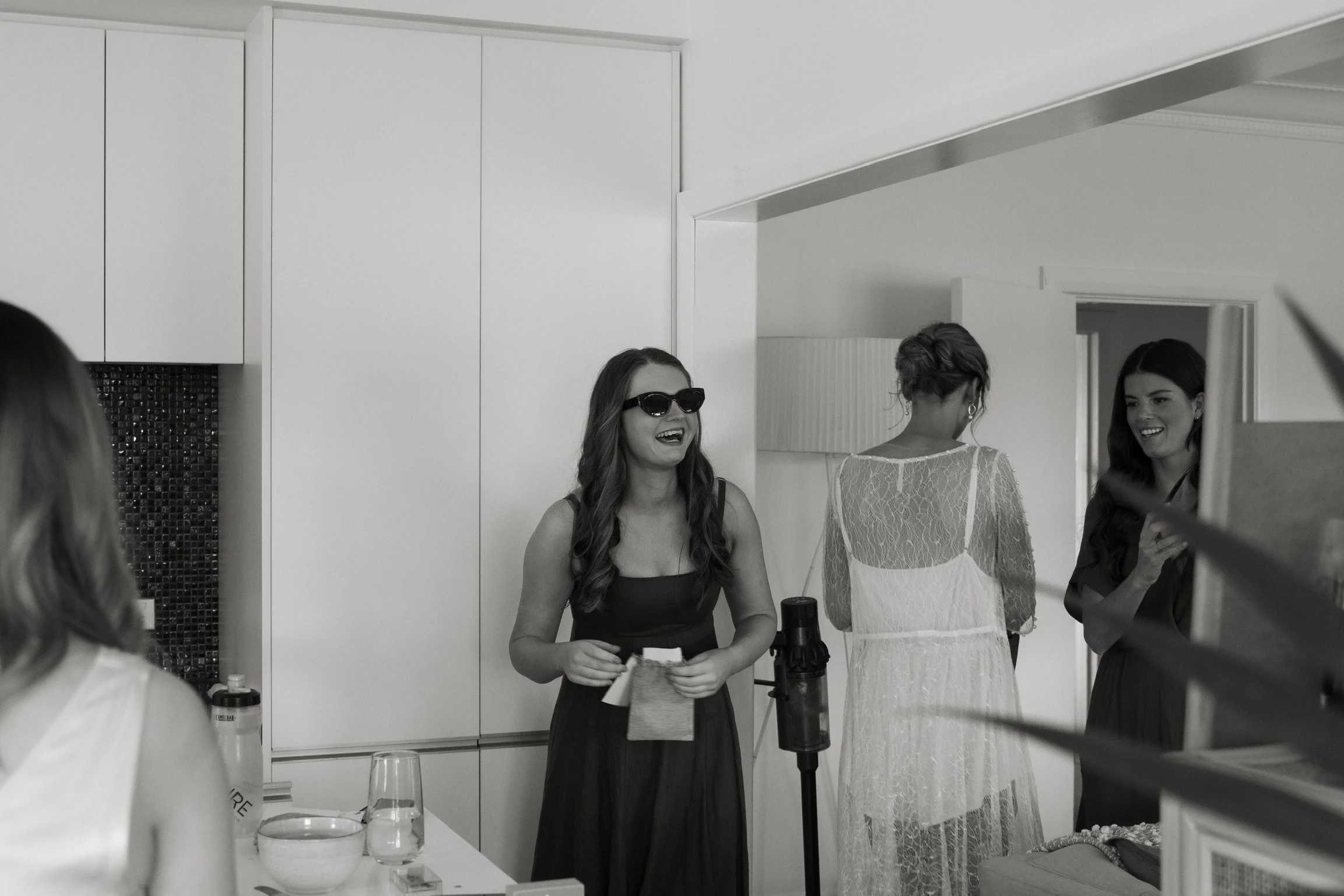 Group of women in a kitchen, one wearing sunglasses and laughing, others chatting, with glasses and bottles on the table.  Wedding Photography Waikato New Zealand. Wedding Photography Hamilton New Zealand. Northland Wedding Photography
