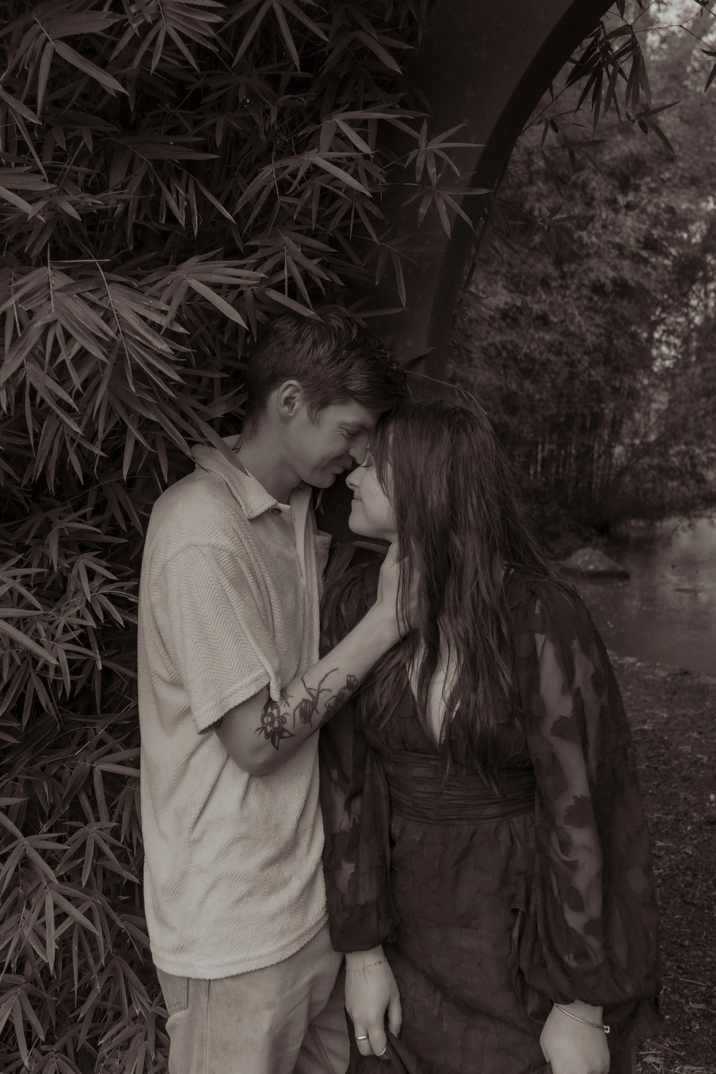 A young man and woman are standing close together outdoors near a bush and a river, touching foreheads and smiling softly. Couples photography Hamilton New Zealand. Engagement photography Hamilton New Zealand. Whangarei Couples photographer