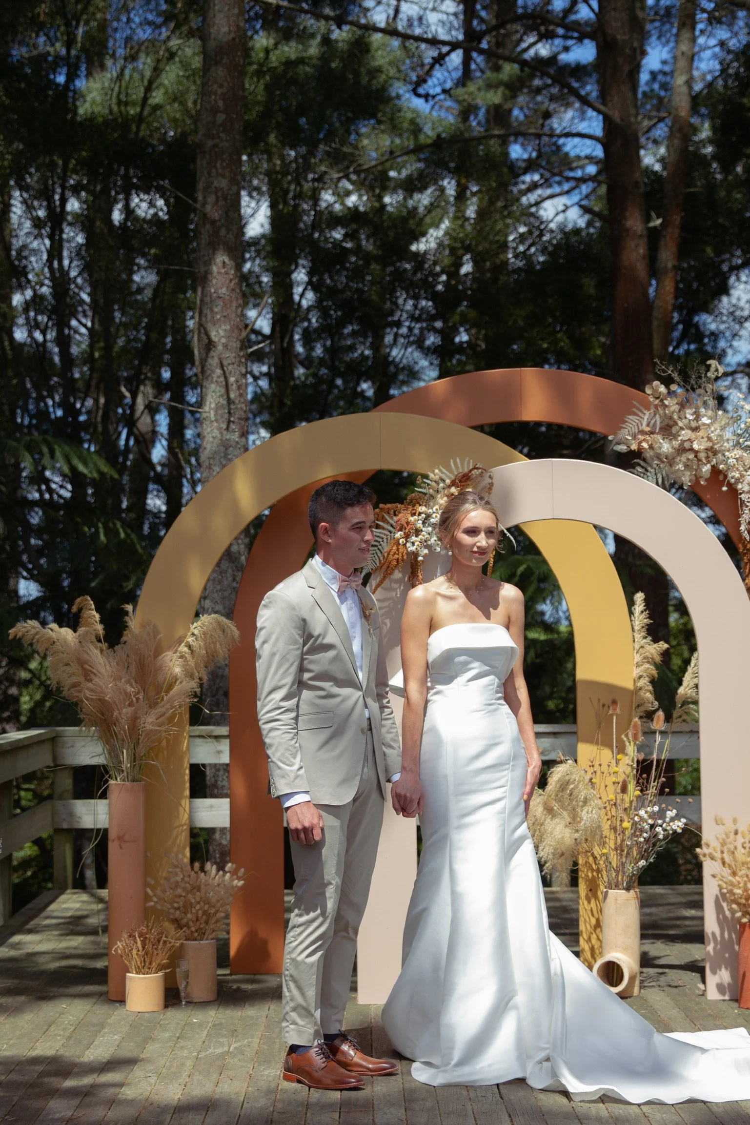 A bride and groom standing outdoors on a wedding altar decorated with dried flowers and large decorative arches, surrounded by trees.  Wedding Photography Waikato New Zealand. Wedding Photography Hamilton New Zealand. Northland Wedding Photography