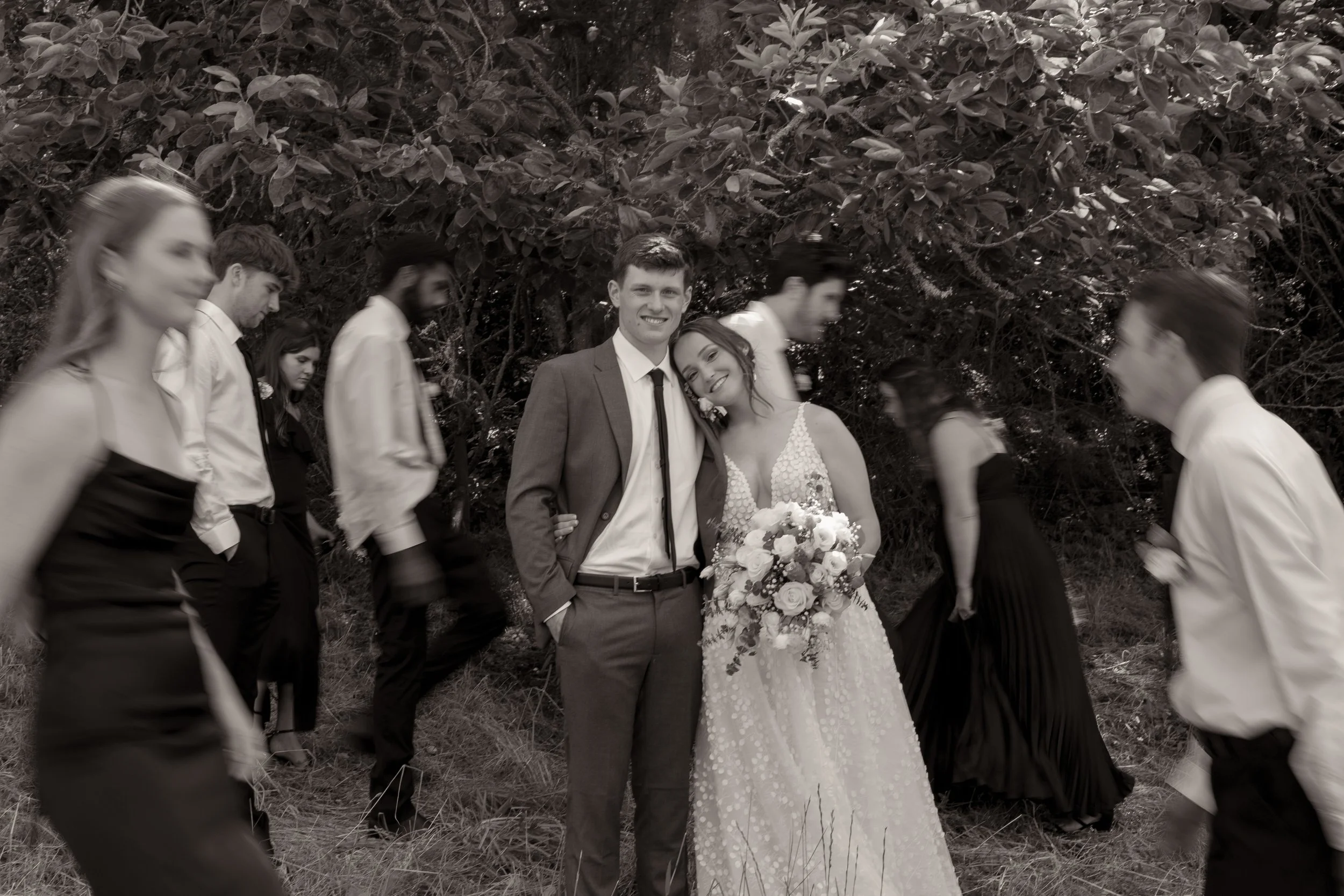Black and white photo of a wedding scene with a smiling bride and groom surrounded by wedding guests in formal attire, walking outdoors with trees in the background.