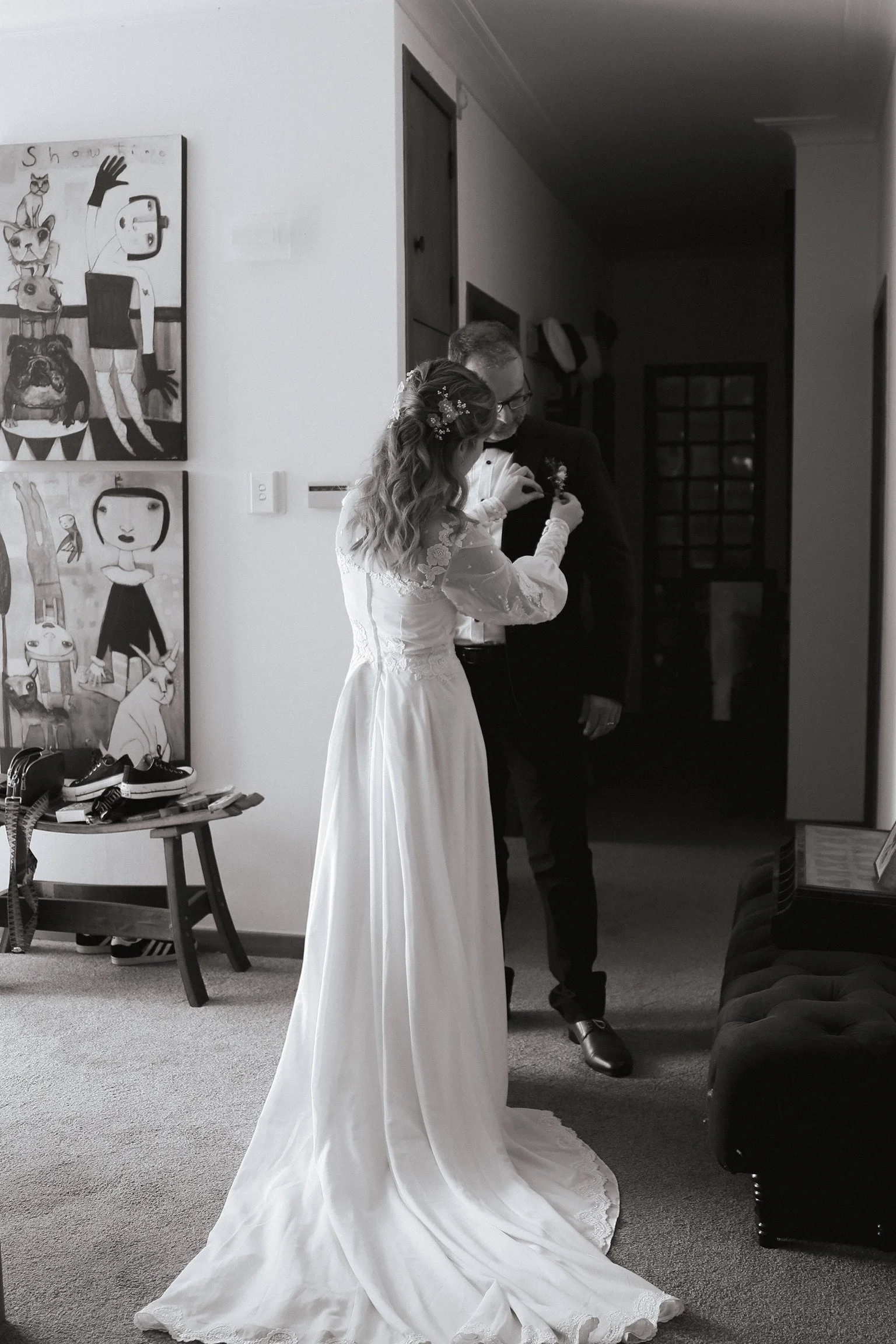 A bride in a wedding dress stands next to a groom in a tuxedo inside a room, preparing for their wedding. The bride has wavy hair with floral hair accessories, and the groom wears glasses. Artwork hangs on the wall behind them. Wedding Photography