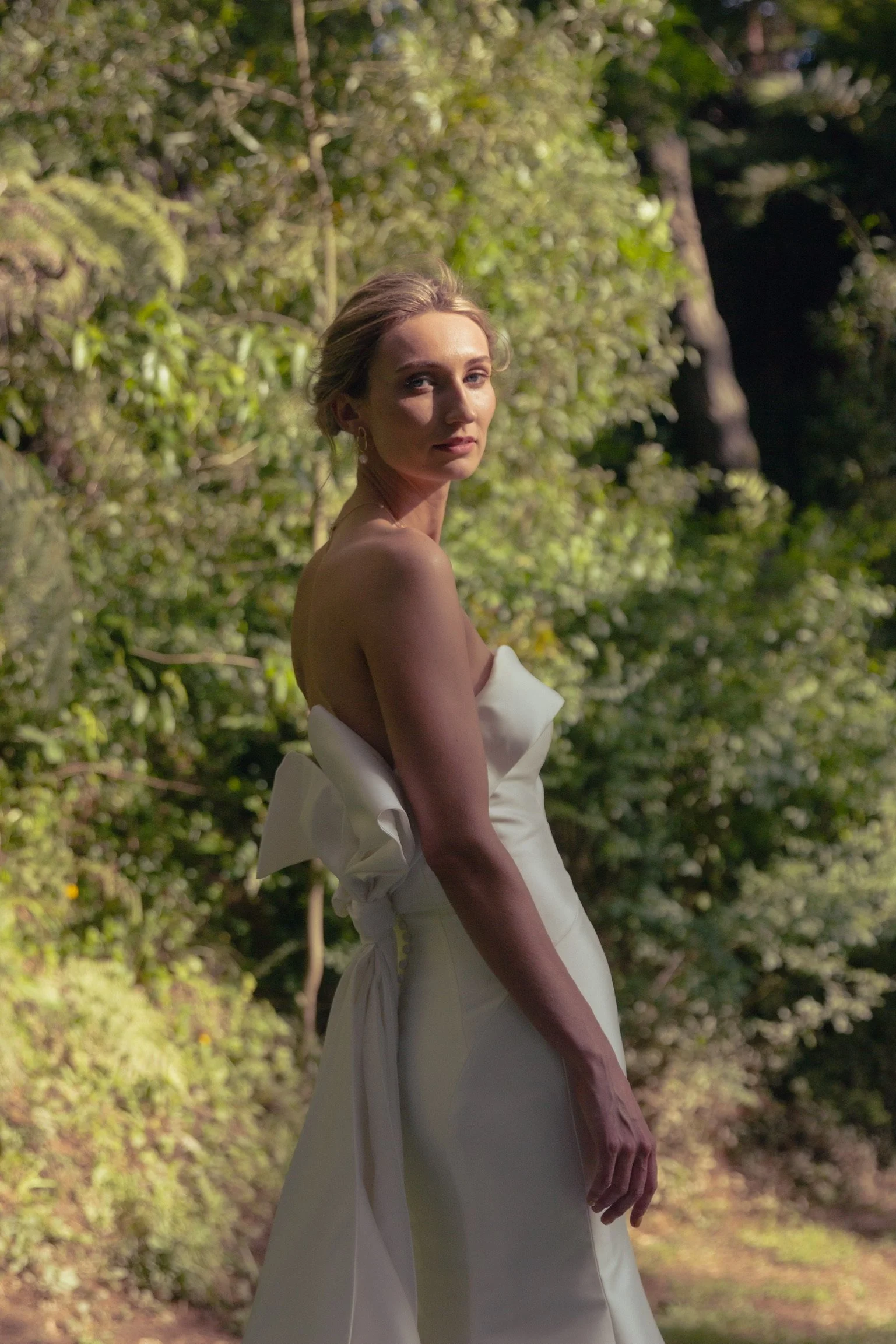 A woman in a strapless white dress stands outdoors against a background of green trees and foliage, looking over her shoulder at the camera.  Wedding Photography Waikato New Zealand. Wedding Photography Hamilton New Zealand. Northland Wedding Photo